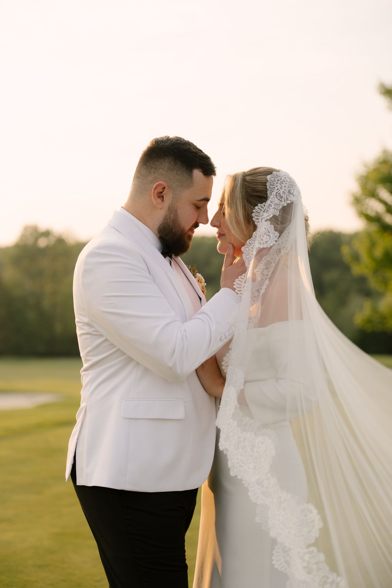 Bride and groom touching foreheads romantically at golden hour, bride in lace veil, groom in white tuxedo.