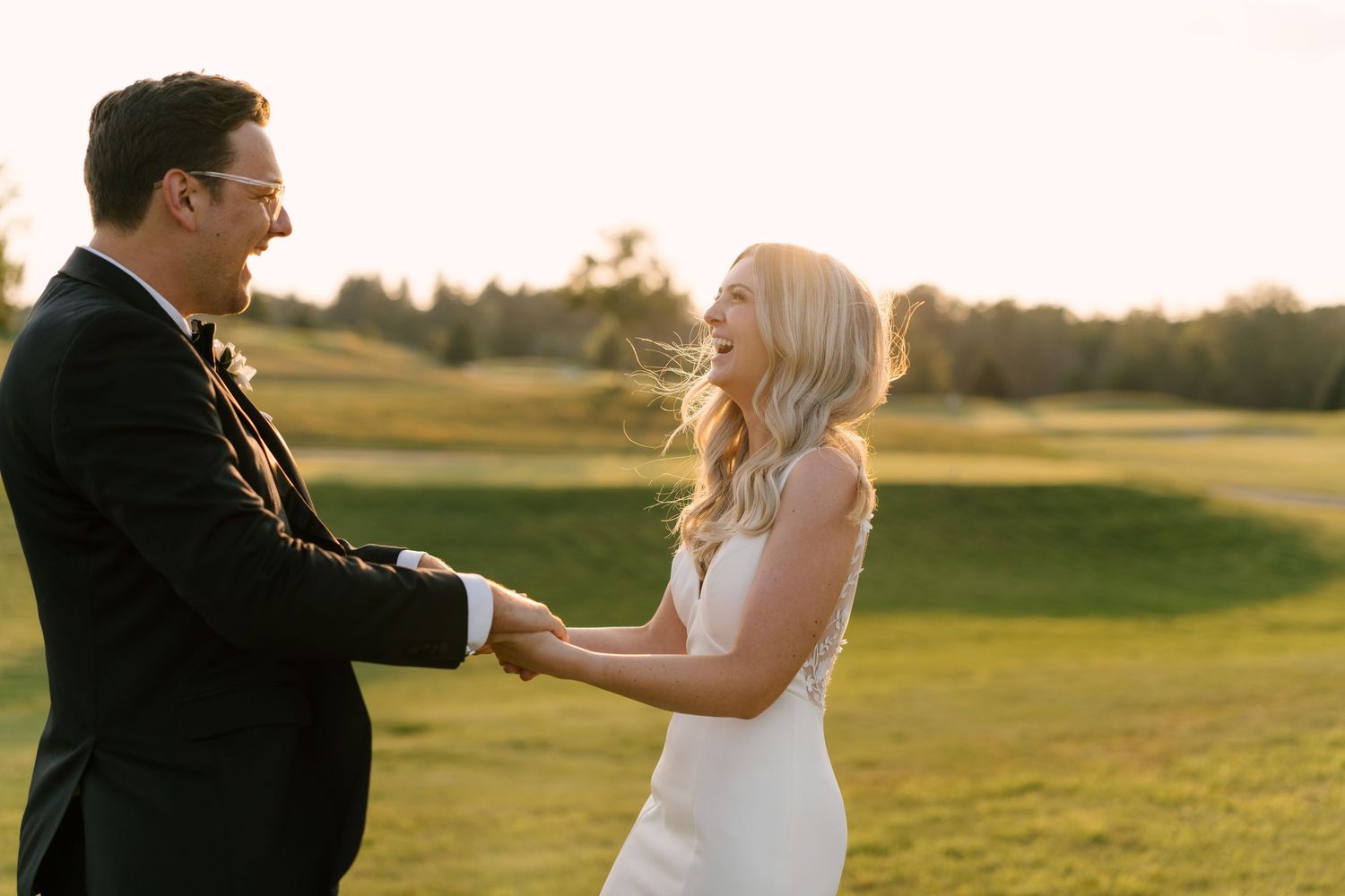 Bride and groom holding hands and laughing together on a sunny green golf course.