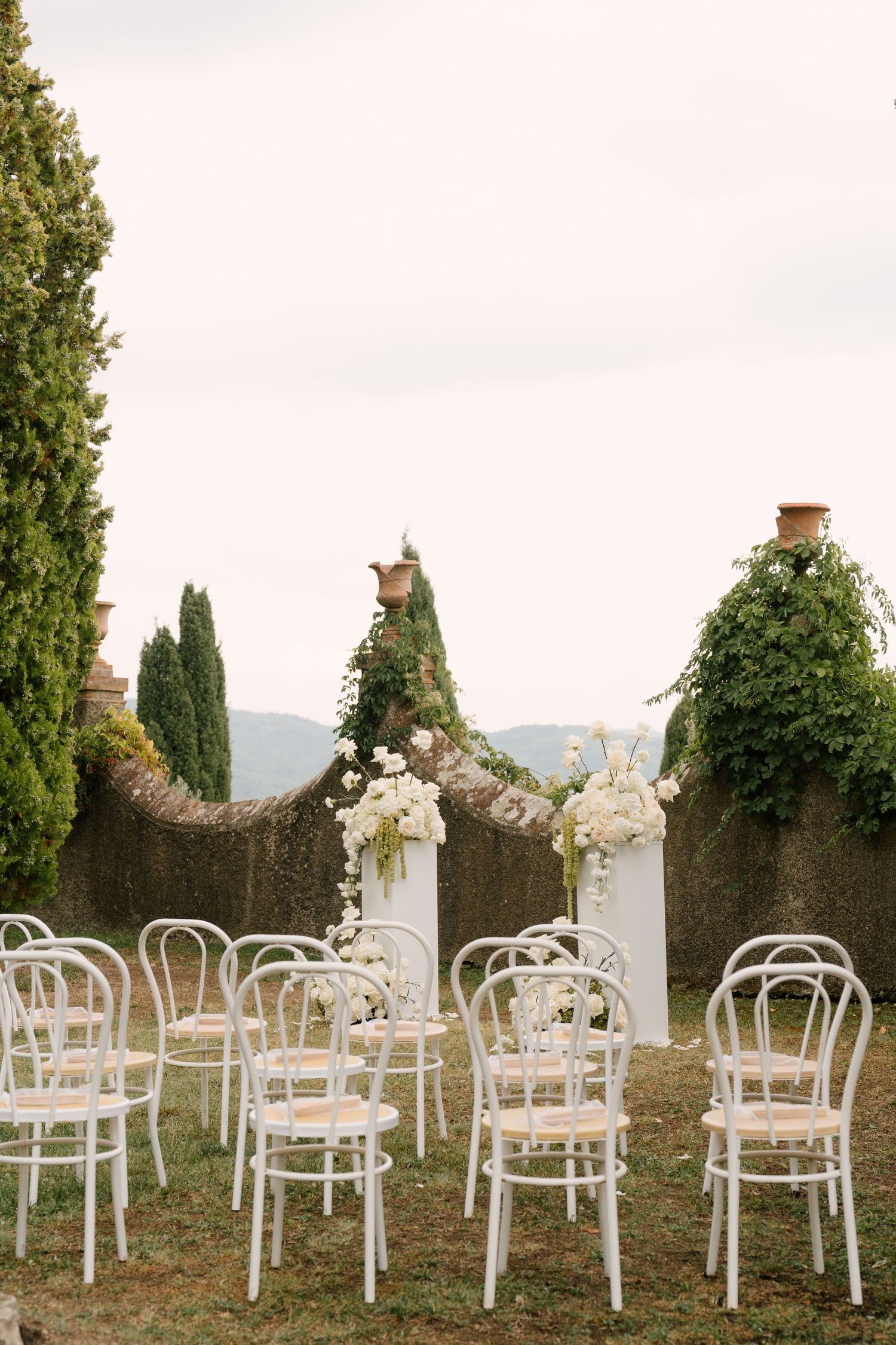 Elegant outdoor wedding ceremony setup with white bistro chairs and floral arrangements against a lush garden backdrop.