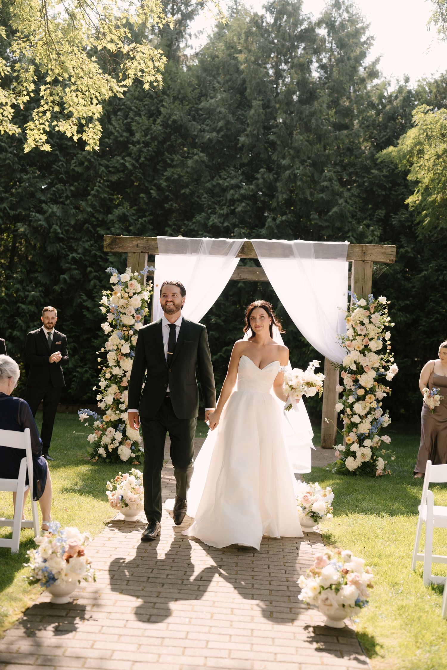 Bride and groom walk hand-in-hand down floral-lined aisle at outdoor wedding ceremony with wooden arch backdrop at Roseville Estates venue.