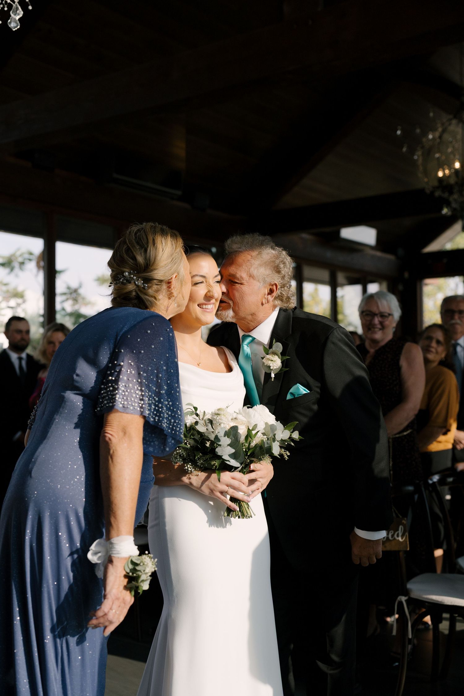 Bride smiling as parents kiss her cheeks during wedding processional in rustic venue with dark wood beams at Cambridge Mill.