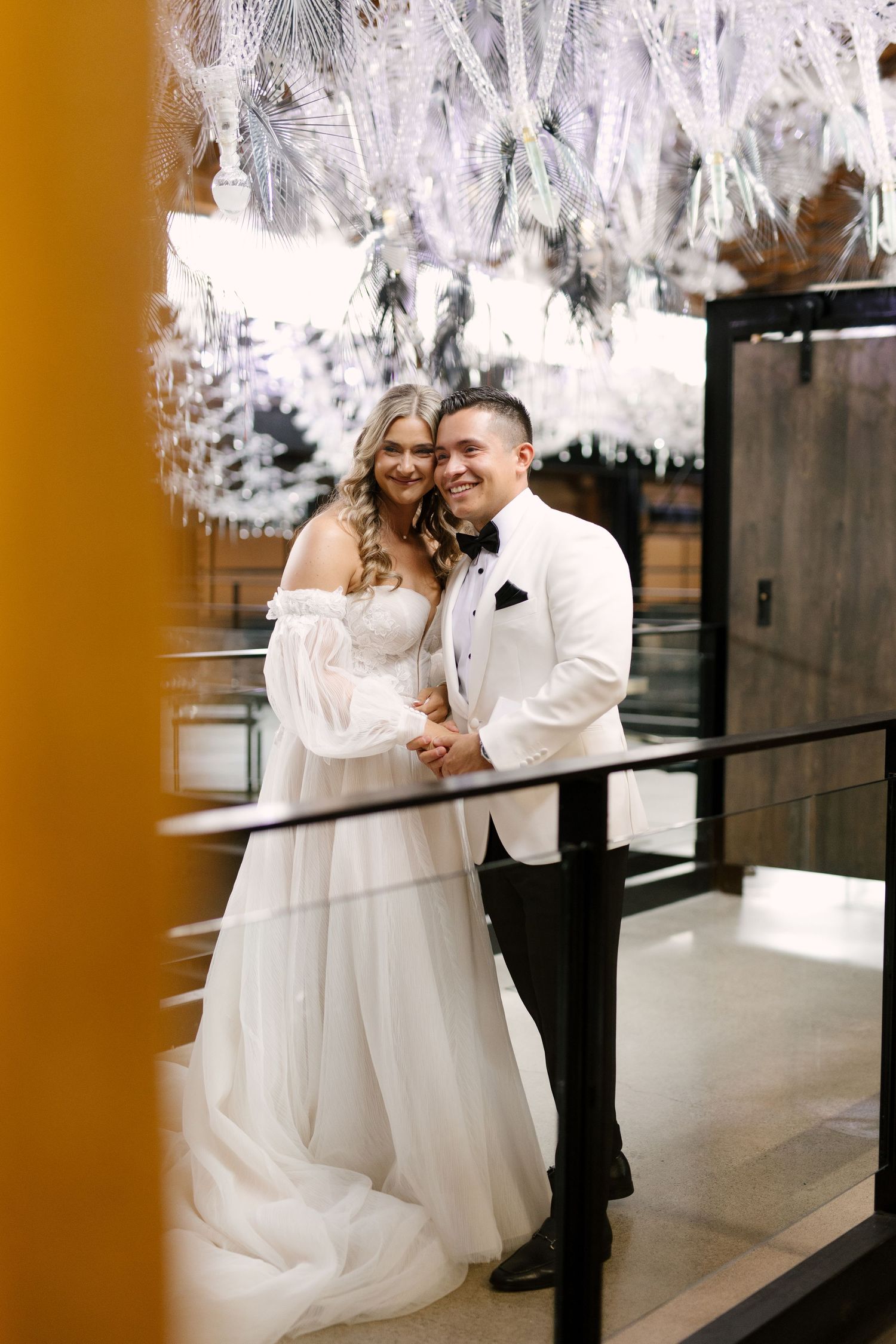 Smiling bride and groom embracing on a balcony beneath white floral chandeliers in an elegant wedding venue.