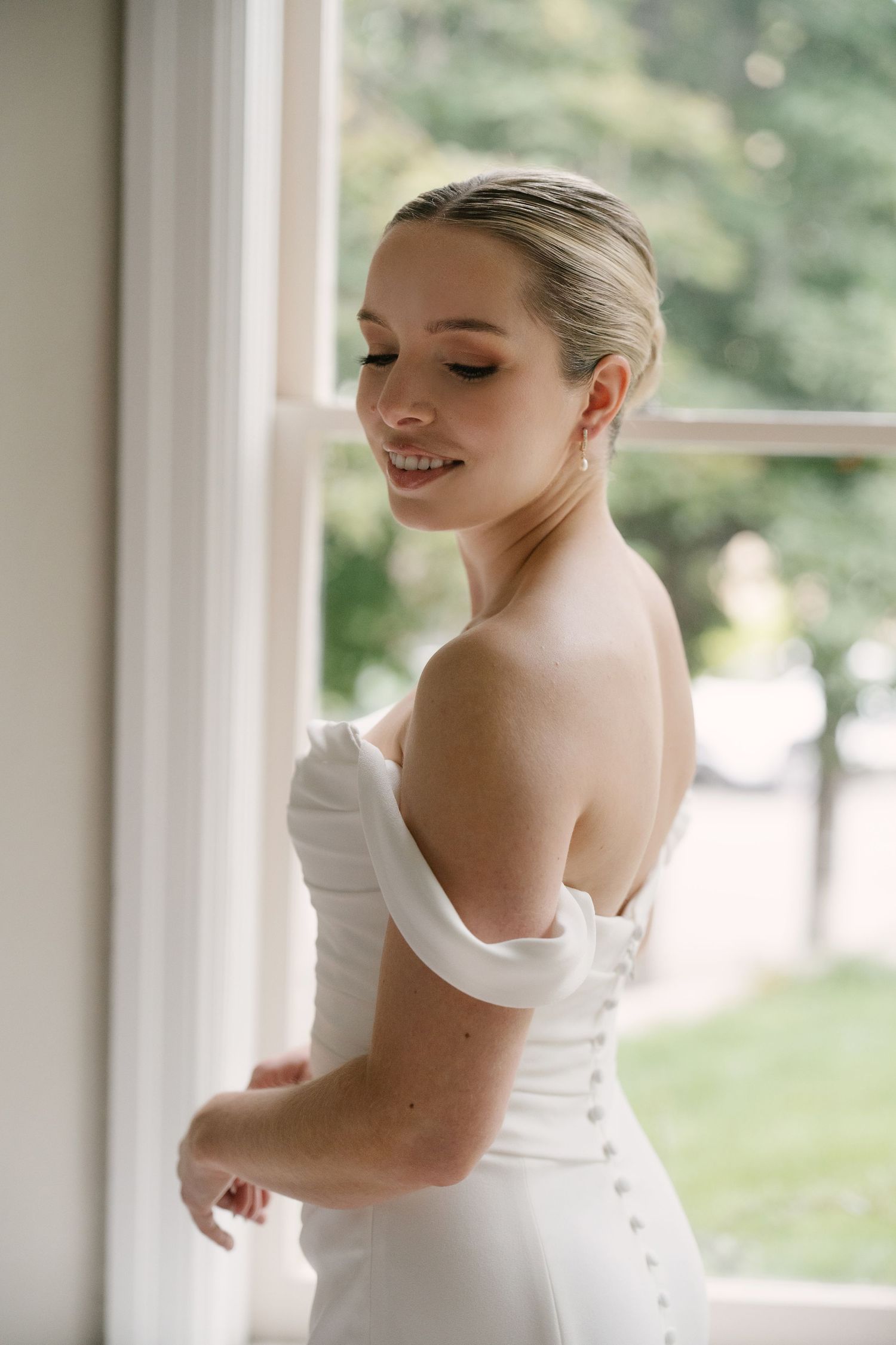 Radiant bride in off-shoulder white wedding dress smiling beside a sunlit window in Cambridge.