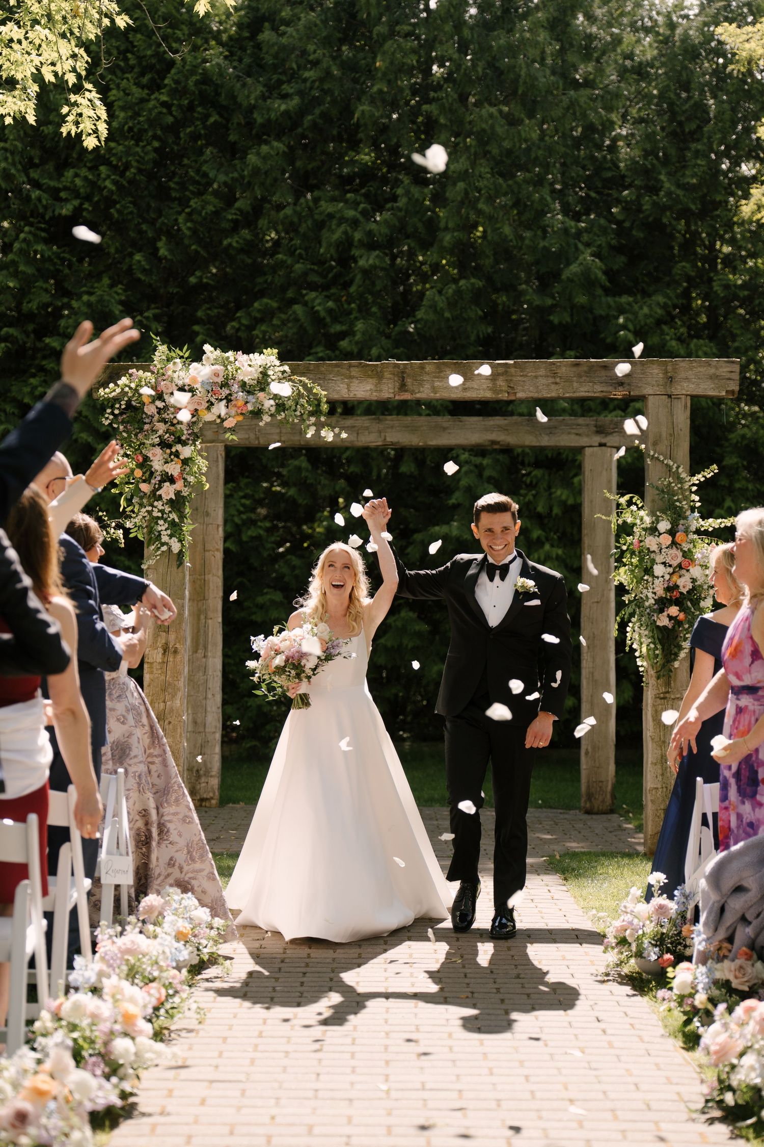 Joyful newlyweds walk down outdoor aisle as guests toss flower petals, bride in white gown holding bouquet.