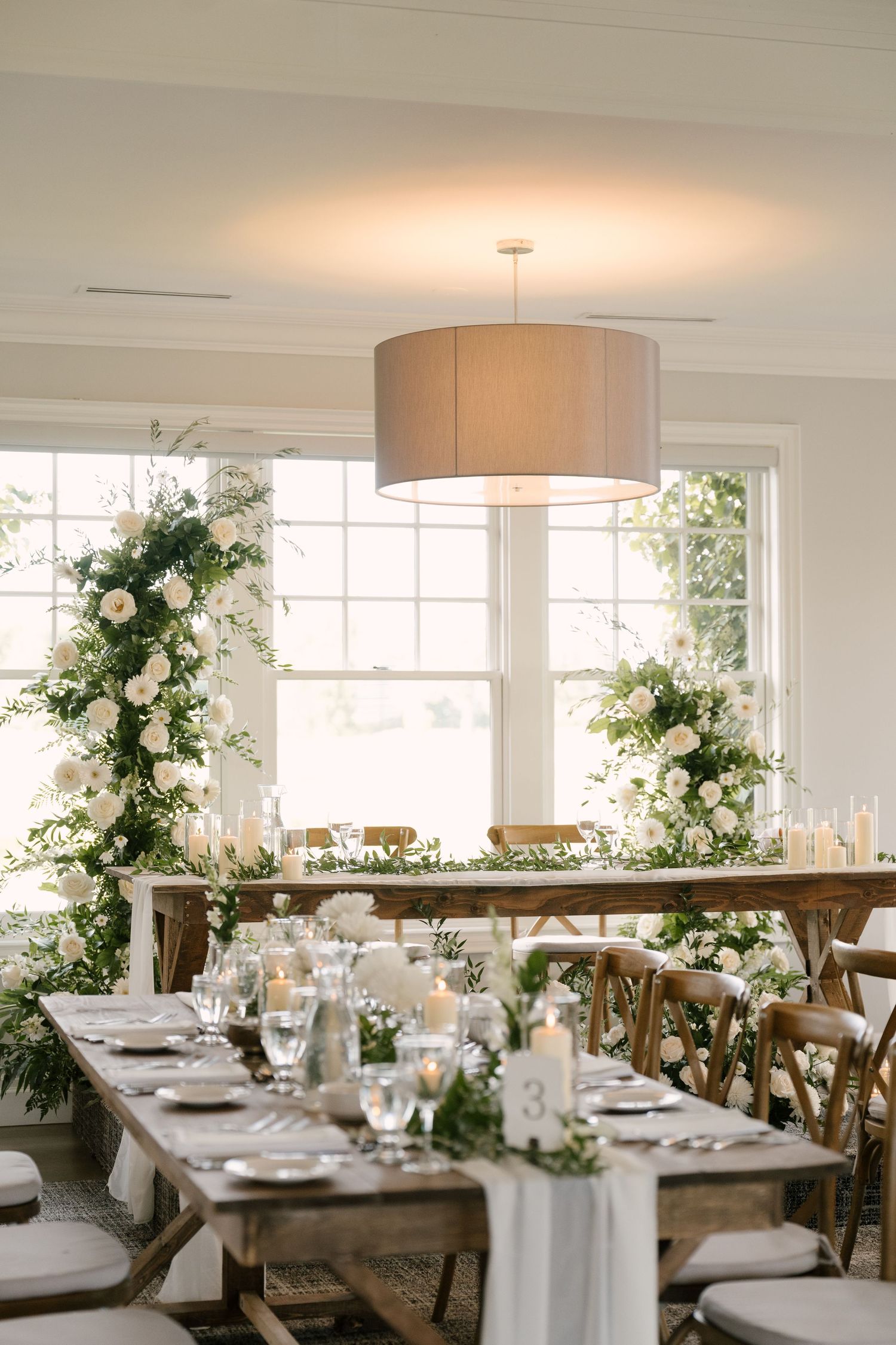 Elegant wedding reception with rustic wood tables, white floral arrangements, candles, and large windows.
