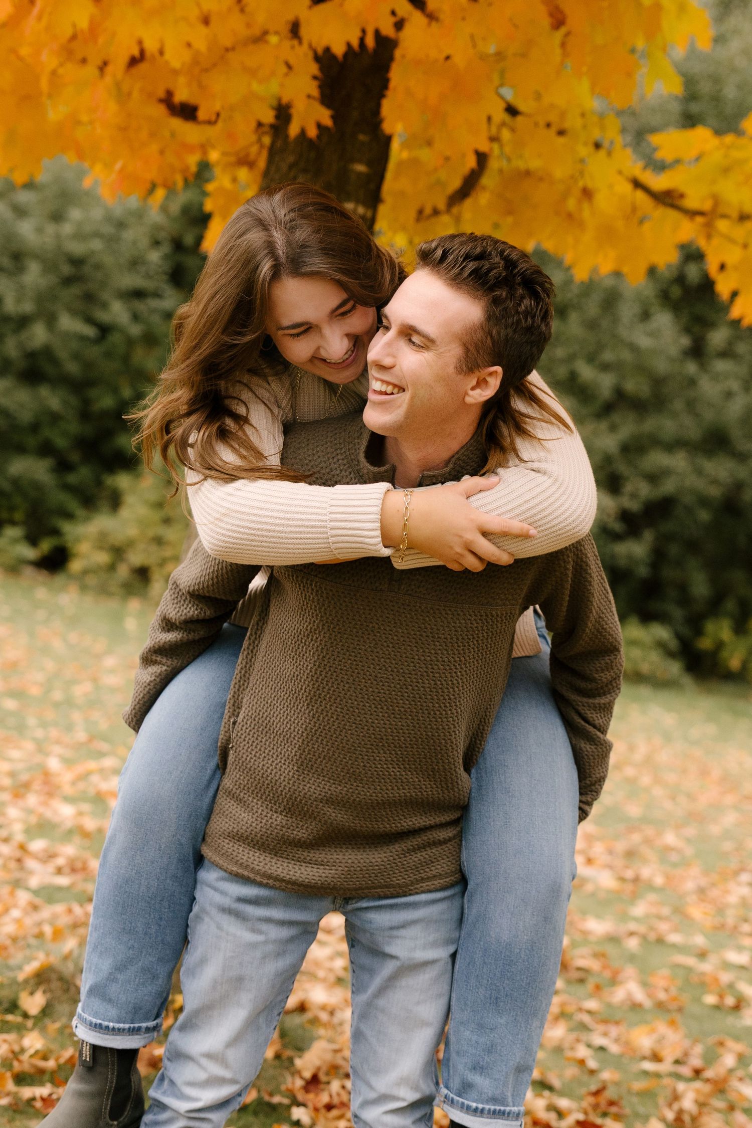 Man giving woman piggyback ride, both laughing, under vibrant yellow maple tree in fall park.