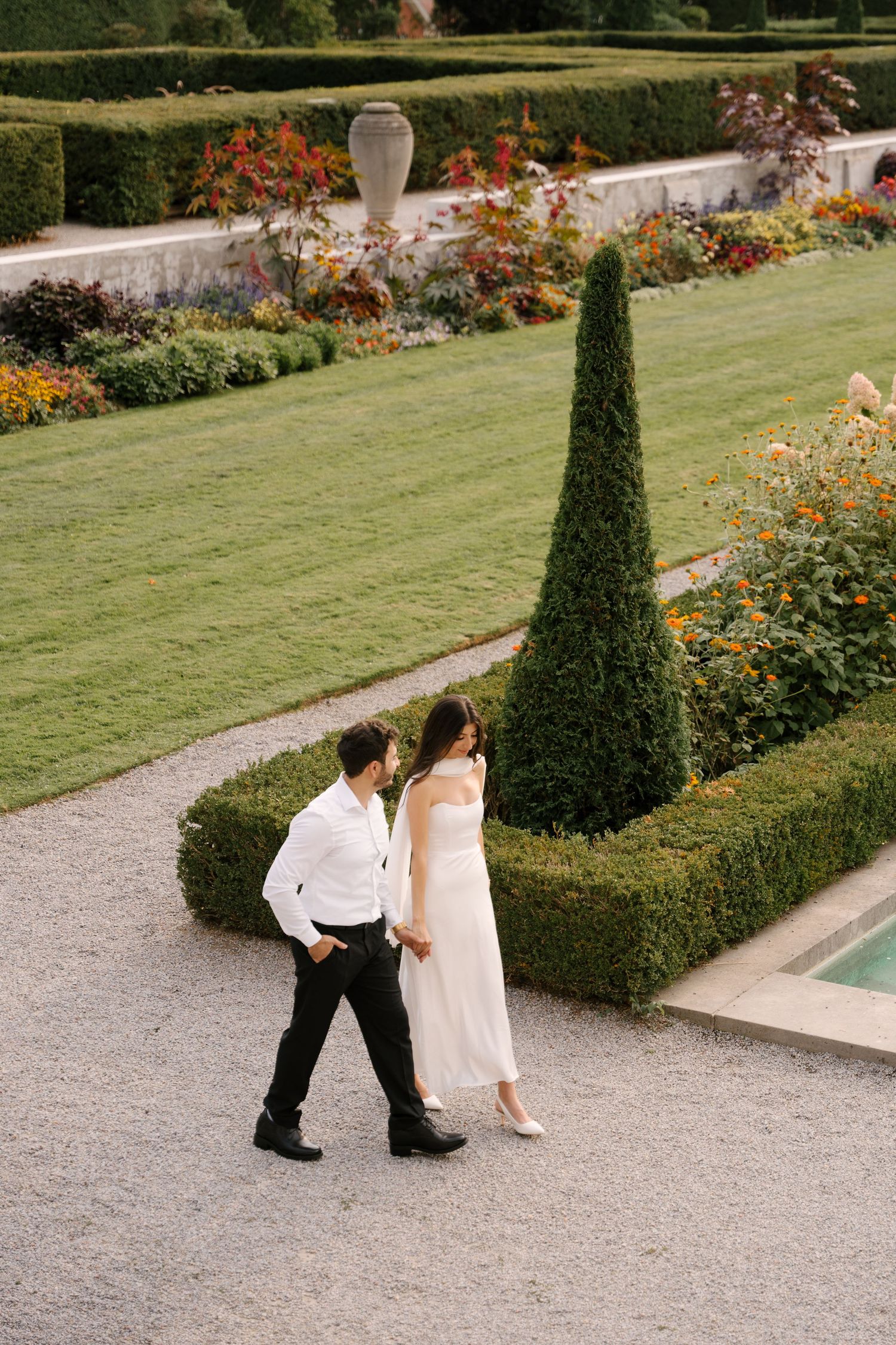 Wedding couple strolling along gravel path beside manicured hedges and colorful flower beds in formal estate garden.