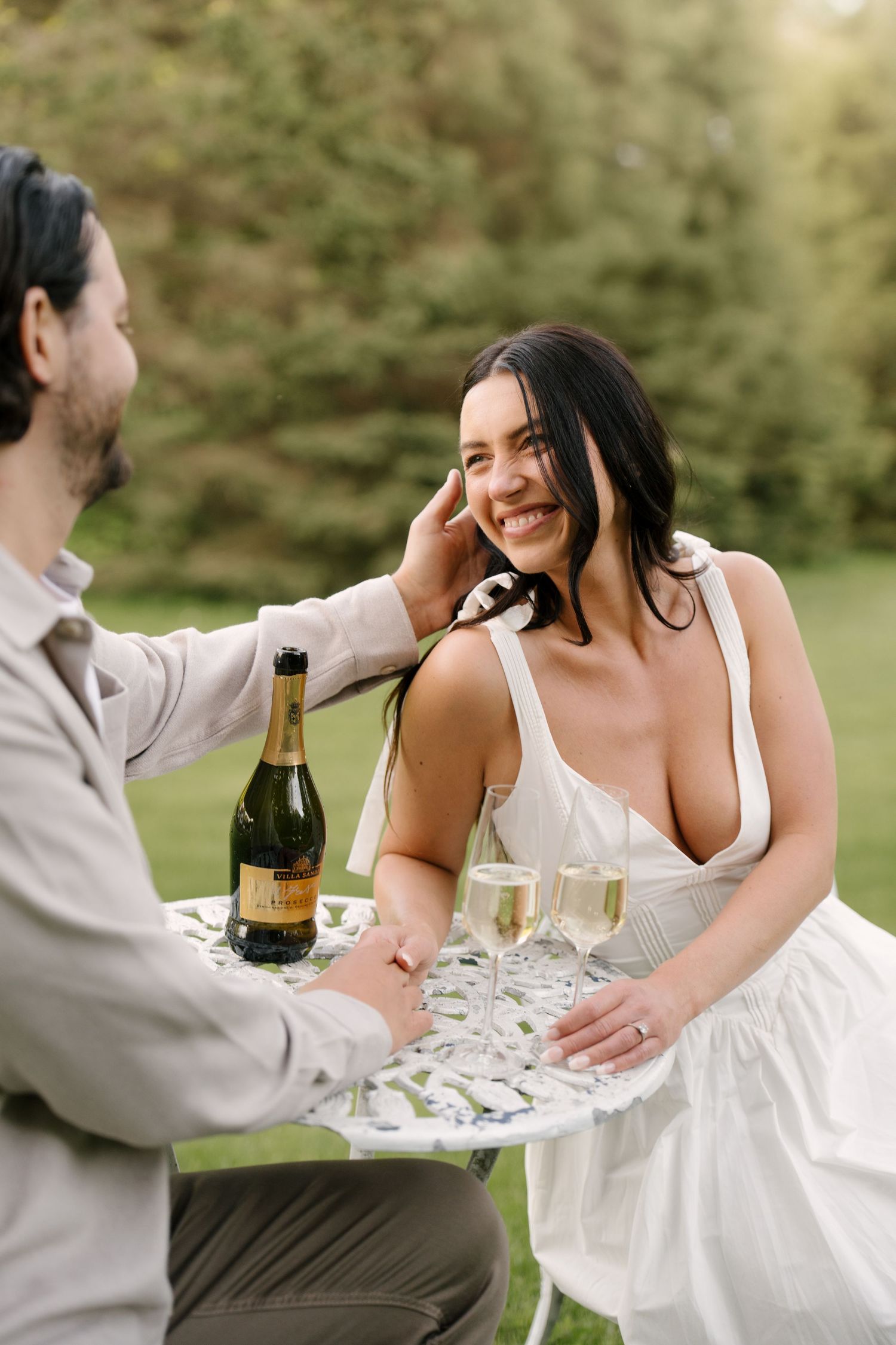 Groom tenderly touching brides face as she smiles at outdoor champagne table celebration.