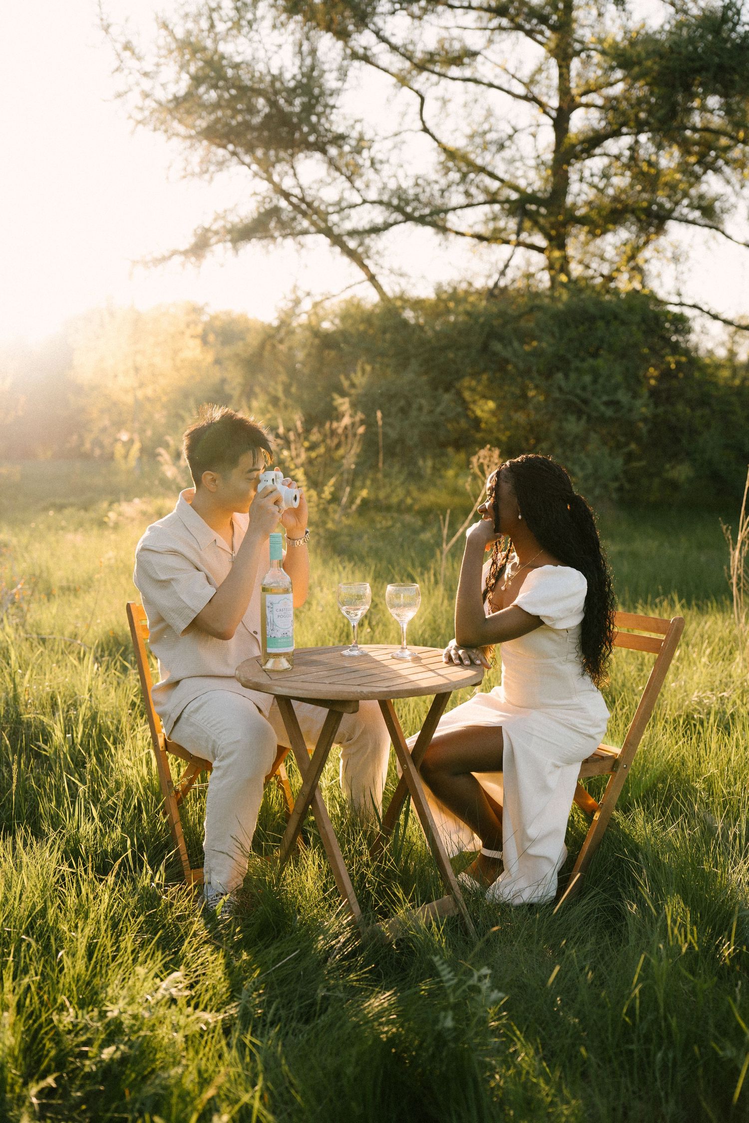 Couple sitting at outdoor table in sunlit green field with wine and camera.