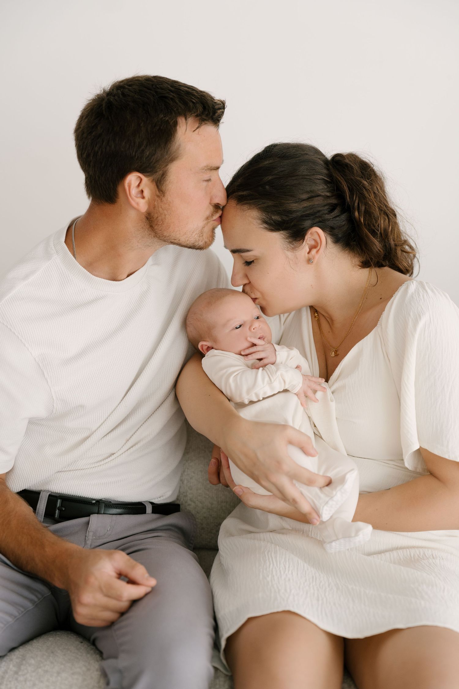 Parents in white outfits kissing their newborn baby while sitting together on couch.