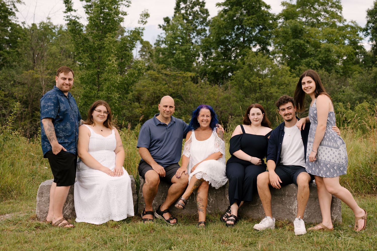 Extended family of seven posing together on large rocks in a lush green outdoor setting.