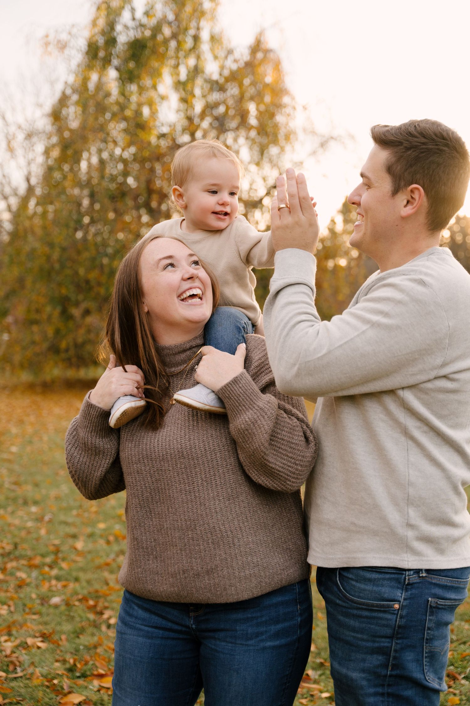 Joyful family with toddler on mom's shoulders high-fiving dad in autumn park.