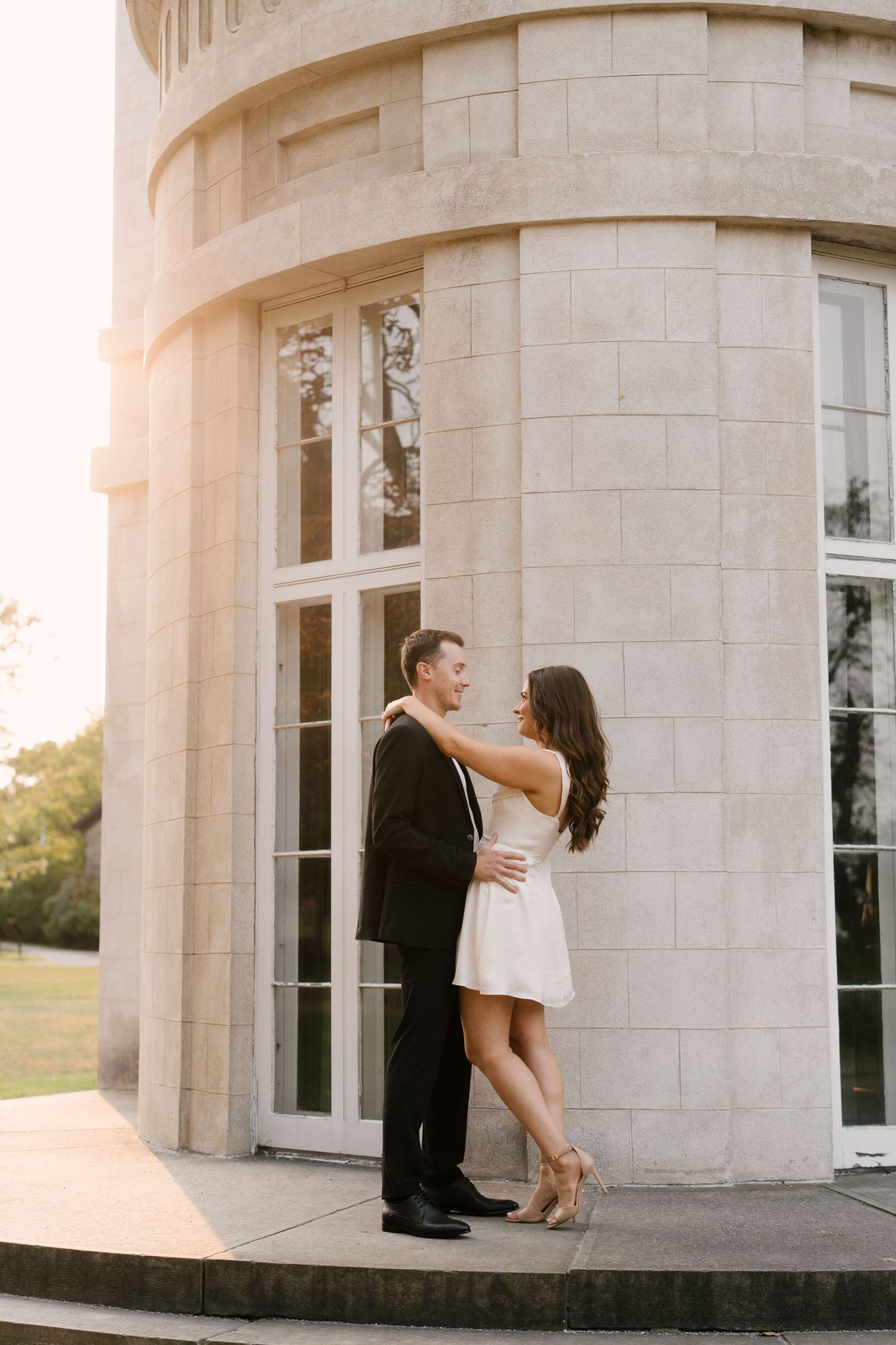 Elegant couple in formal attire embracing beside a stone building at golden hour.
