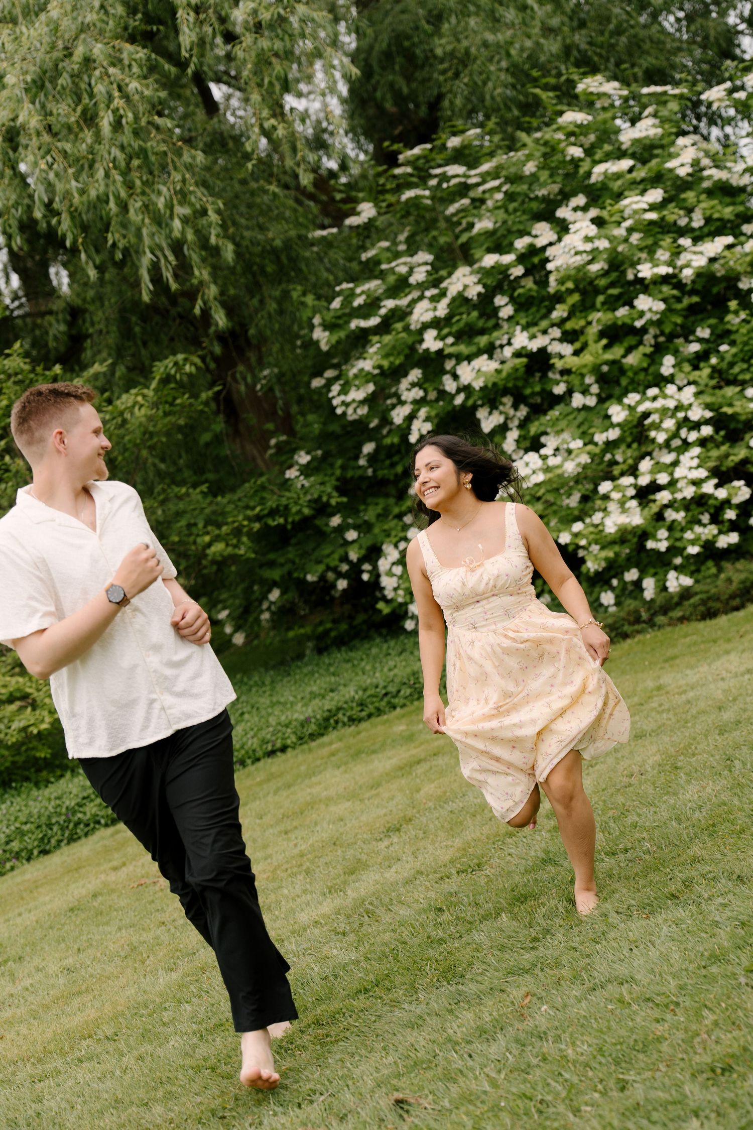 Joyful couple running barefoot through green grass near white blooming trees in a sunny garden.