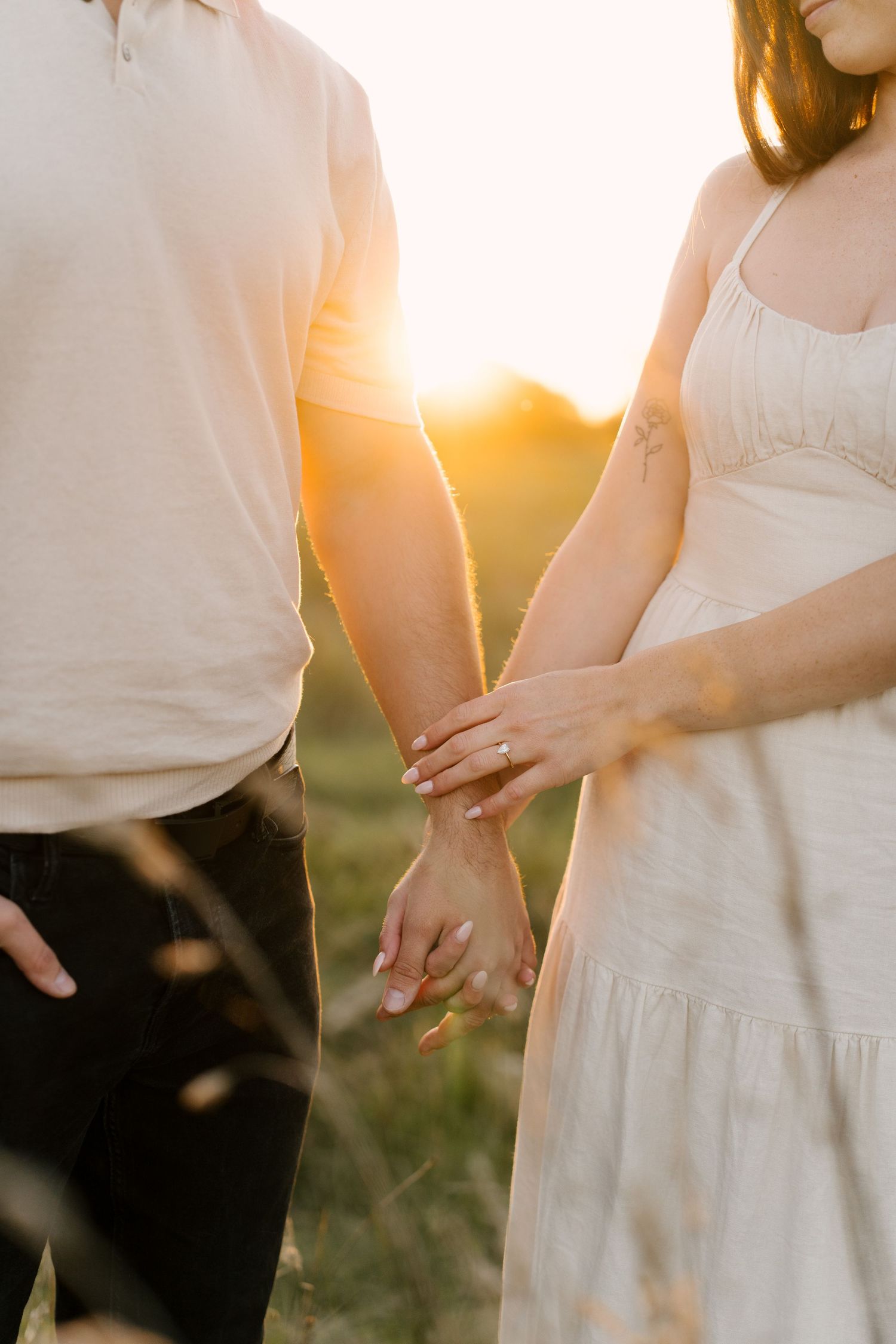 Close-up of couple holding hands with a sparkling engagement ring in a sunset field.