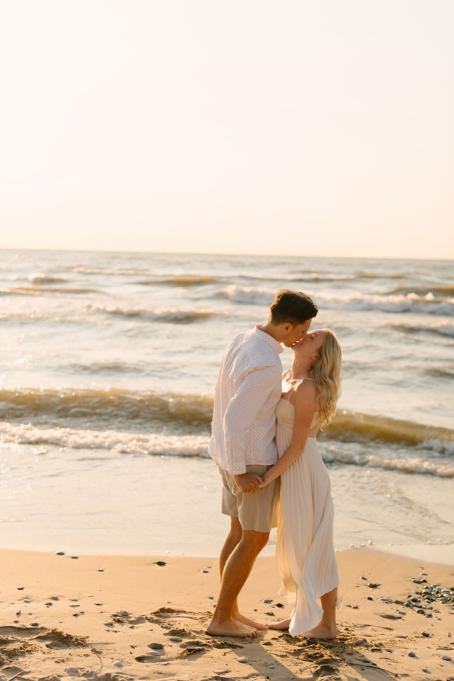 Couple sharing a romantic kiss on a sandy beach at golden sunset with ocean waves in the background.