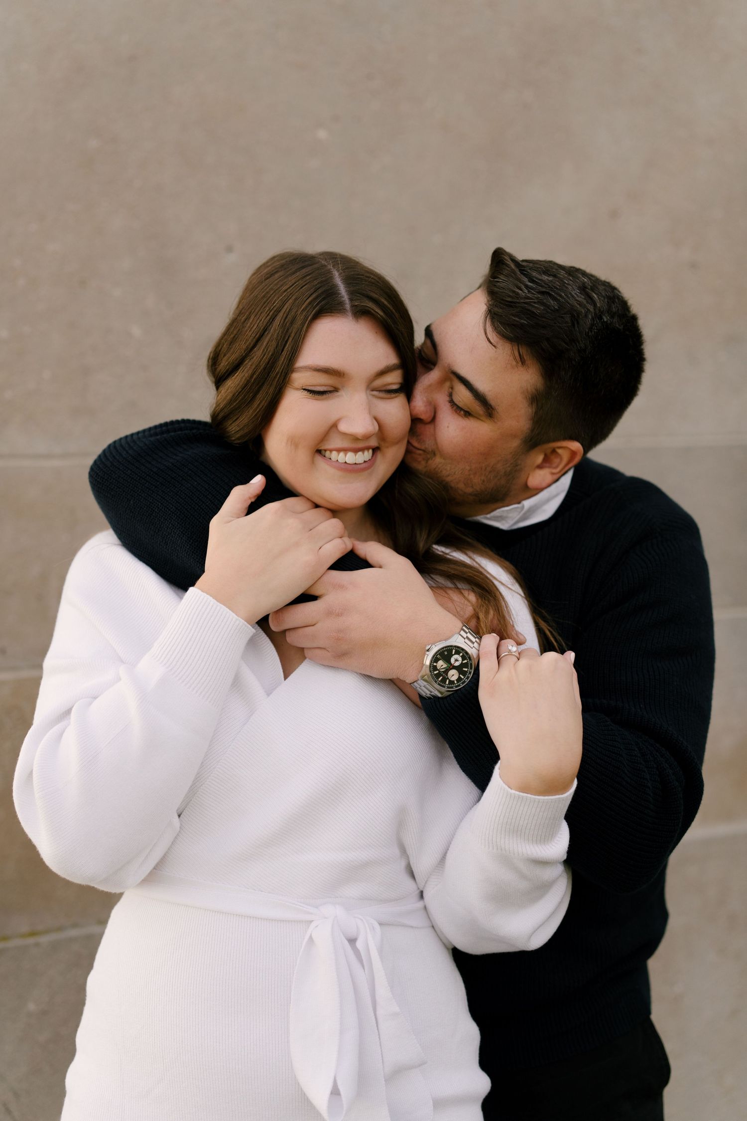 Man in black sweater embracing and kissing woman in white dress from behind.
