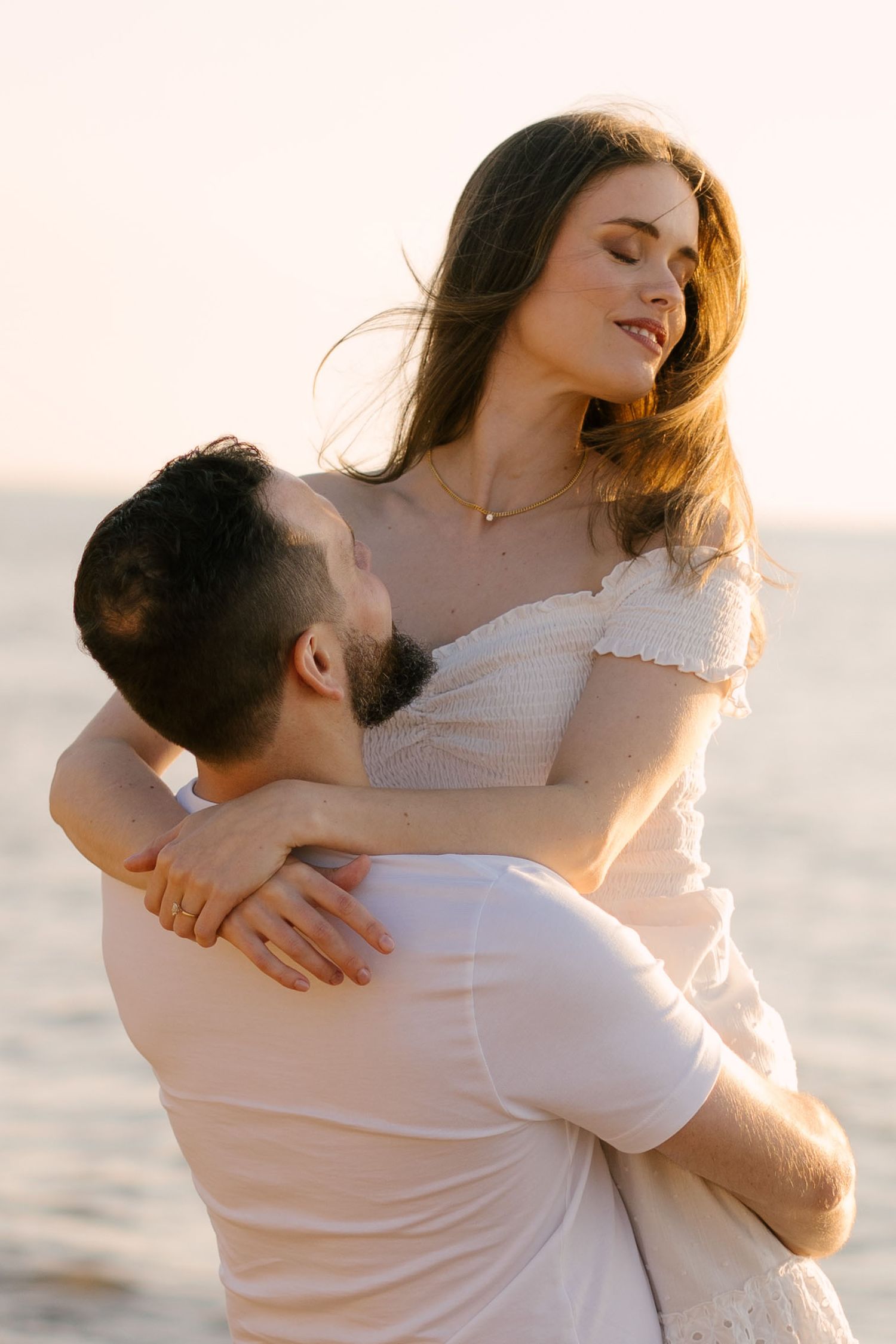 Couple embracing at sunset on the beach, man lifting woman in white dress as golden light reflects off the sea.