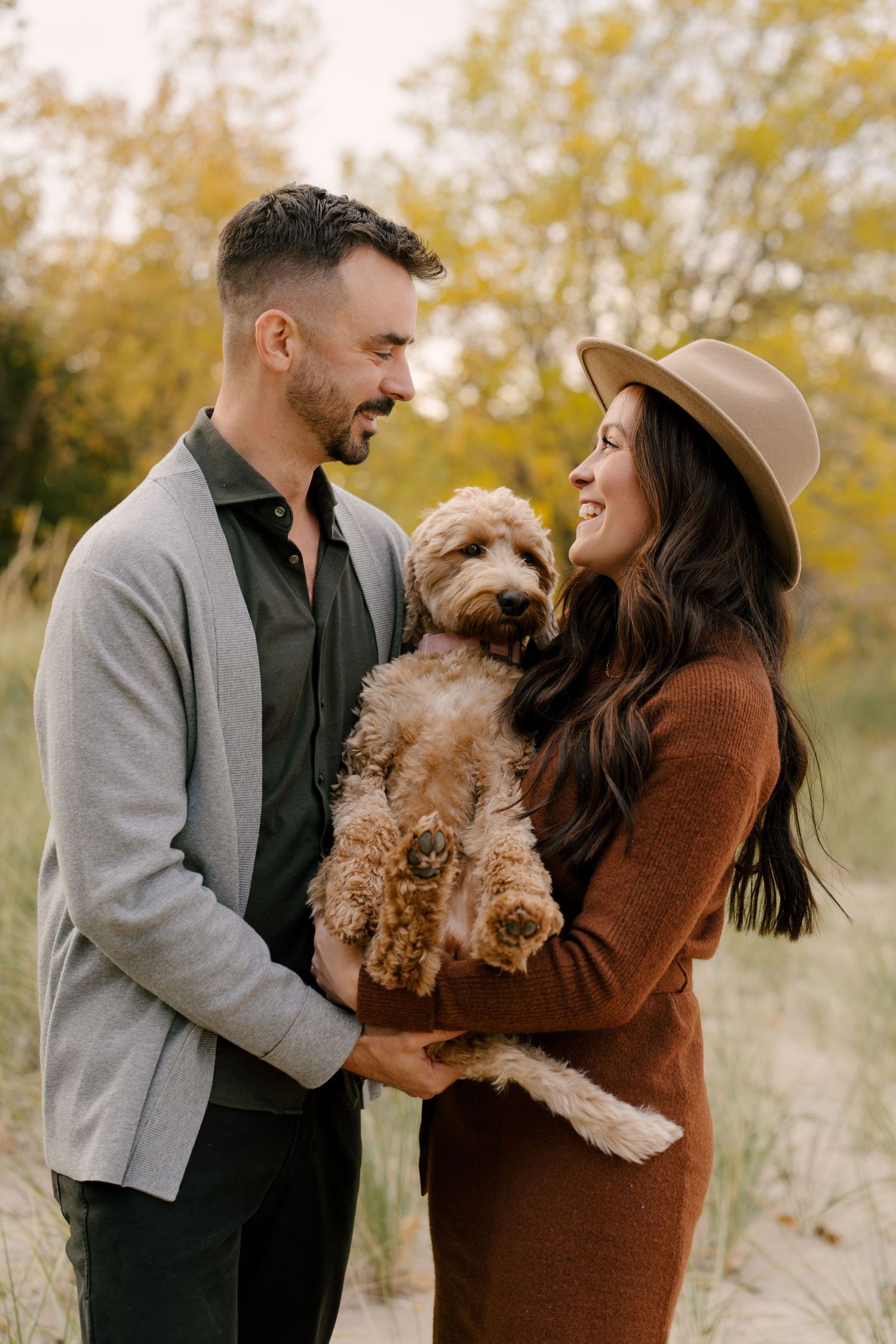 Smiling couple holding fluffy goldendoodle dog outdoors in autumn fall setting.