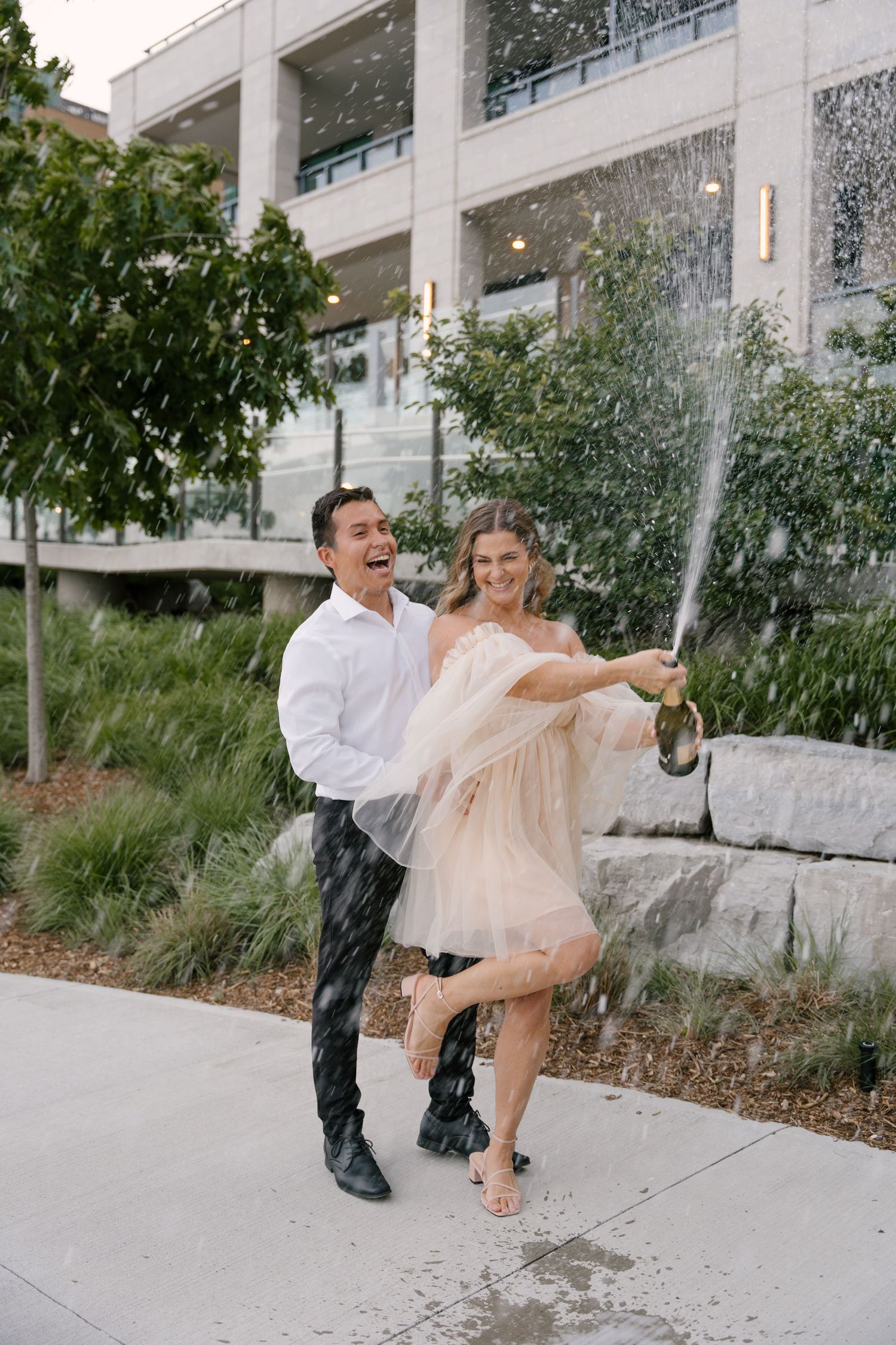 Laughing couple spraying champagne outside a modern building, woman in a blush tulle mini dress.