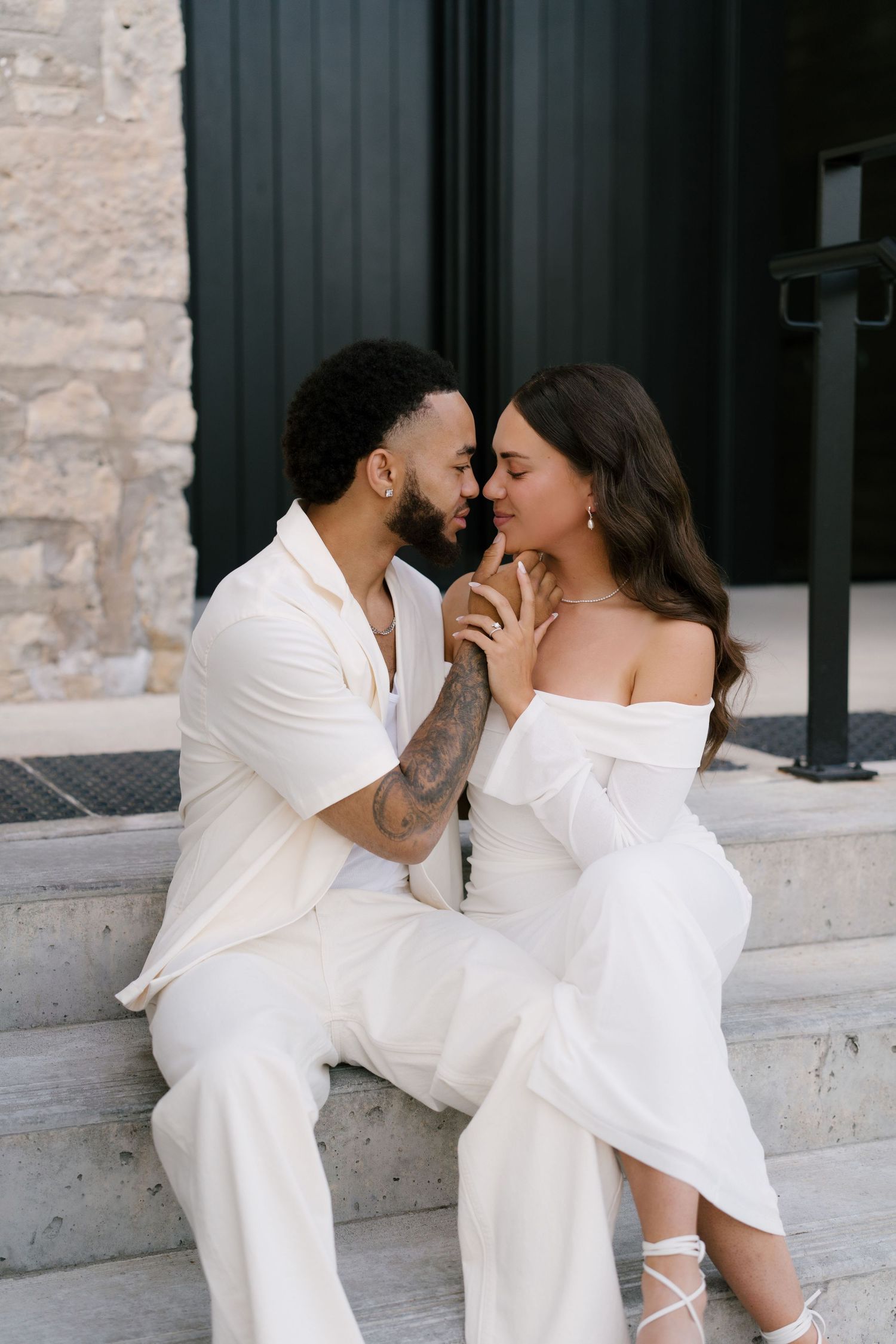 Engaged couple in white outfits sit on steps, leaning in for a kiss while holding hands.