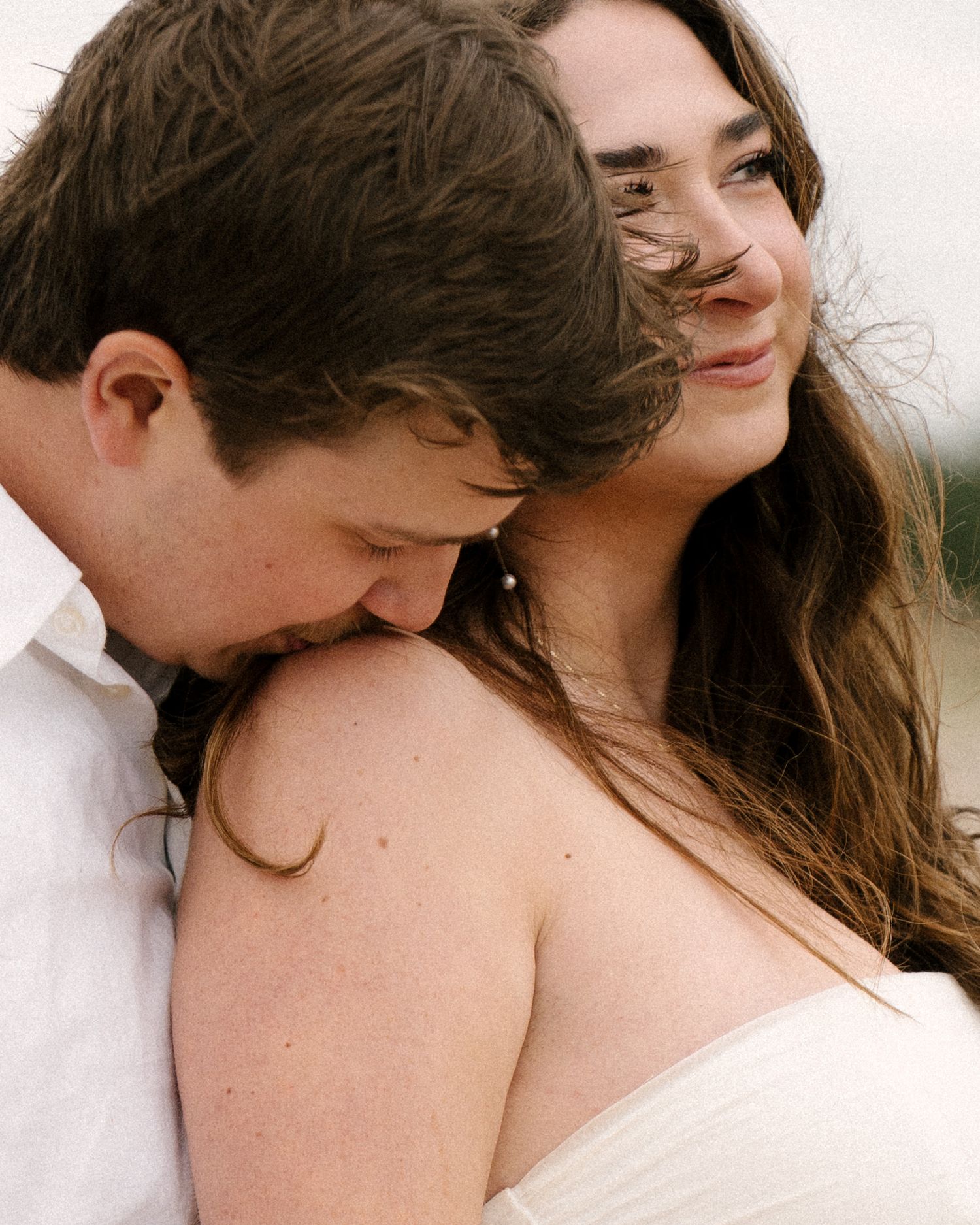 Groom kissing bride's shoulder while she smiles warmly during a windy beach engagement session.