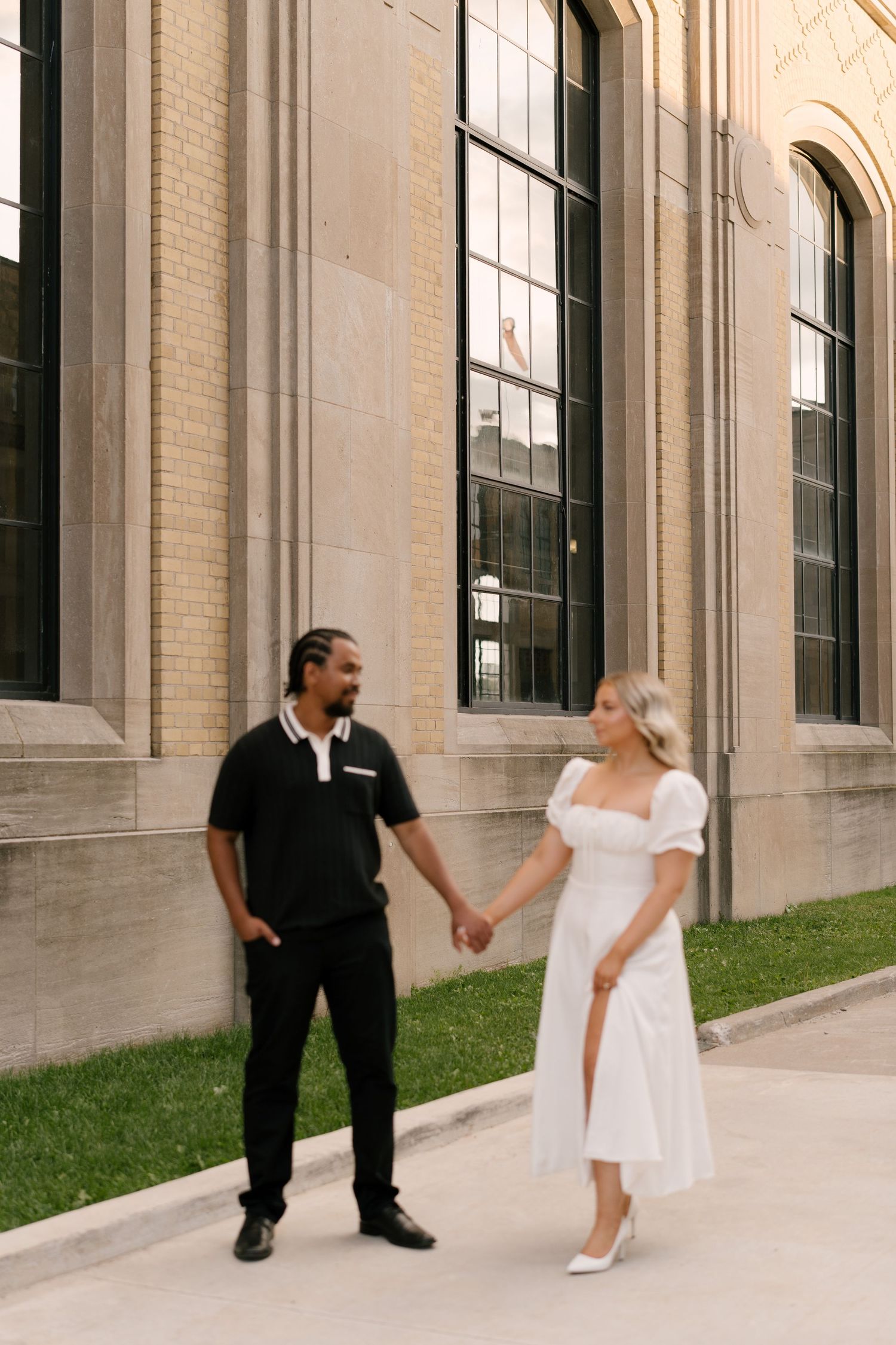Interracial couple holding hands and smiling at each other beside a grand stone building exterior.