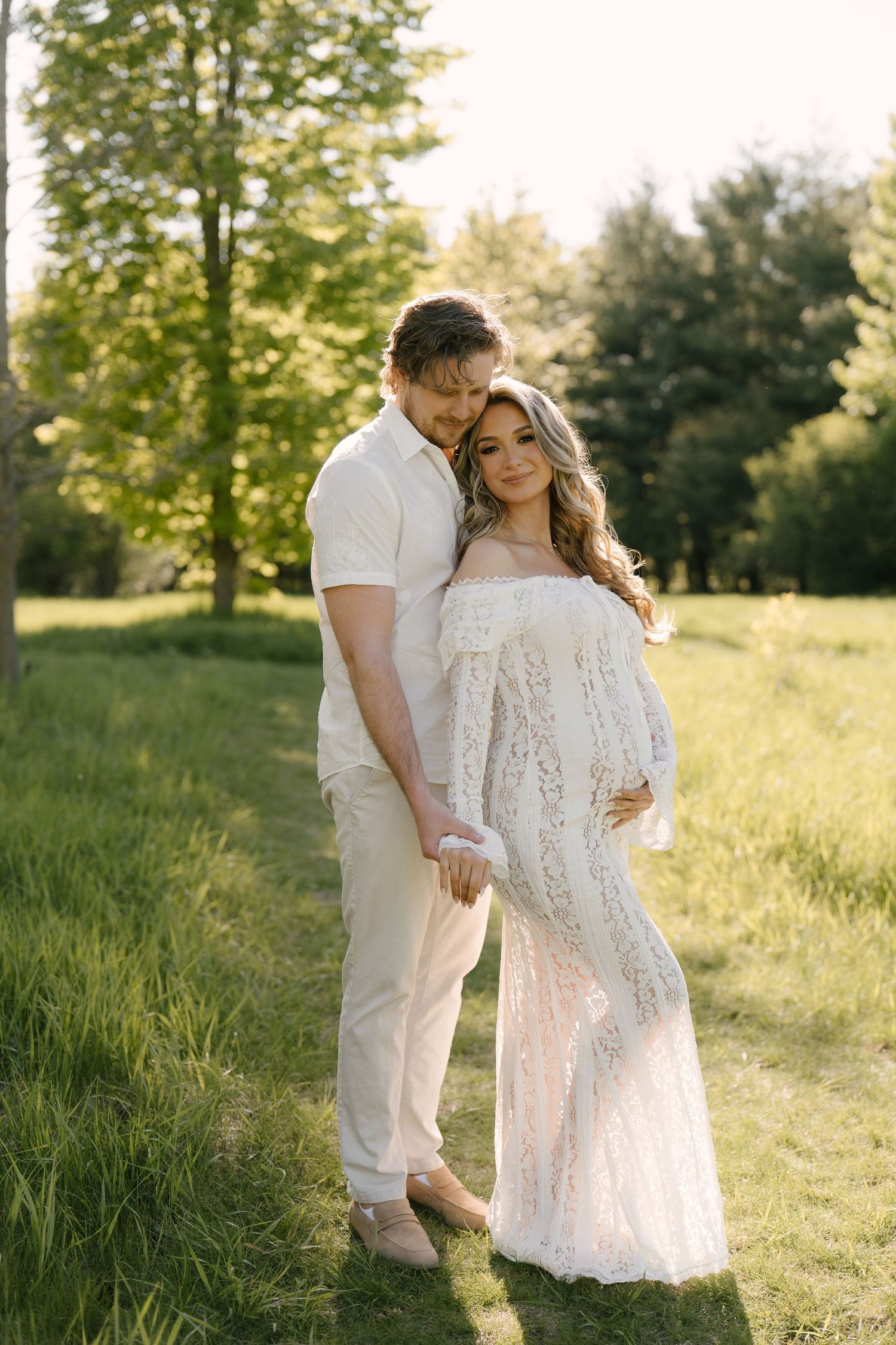 Couple embracing in sunlit park, woman in white lace maternity gown cradling pregnant belly.