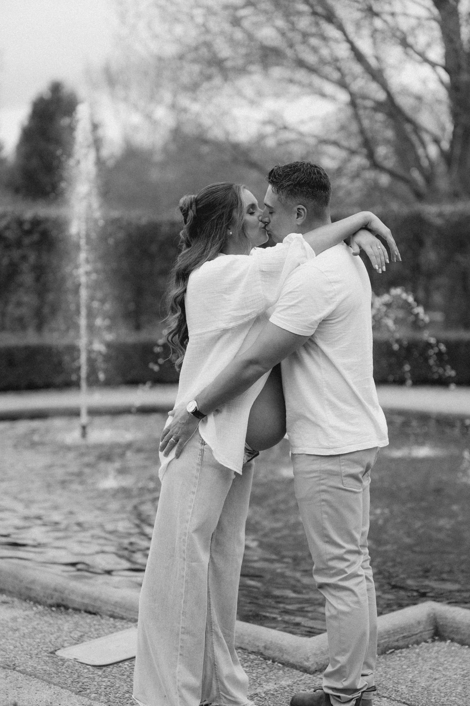 Black and white photo of couple kissing near decorative garden fountain.