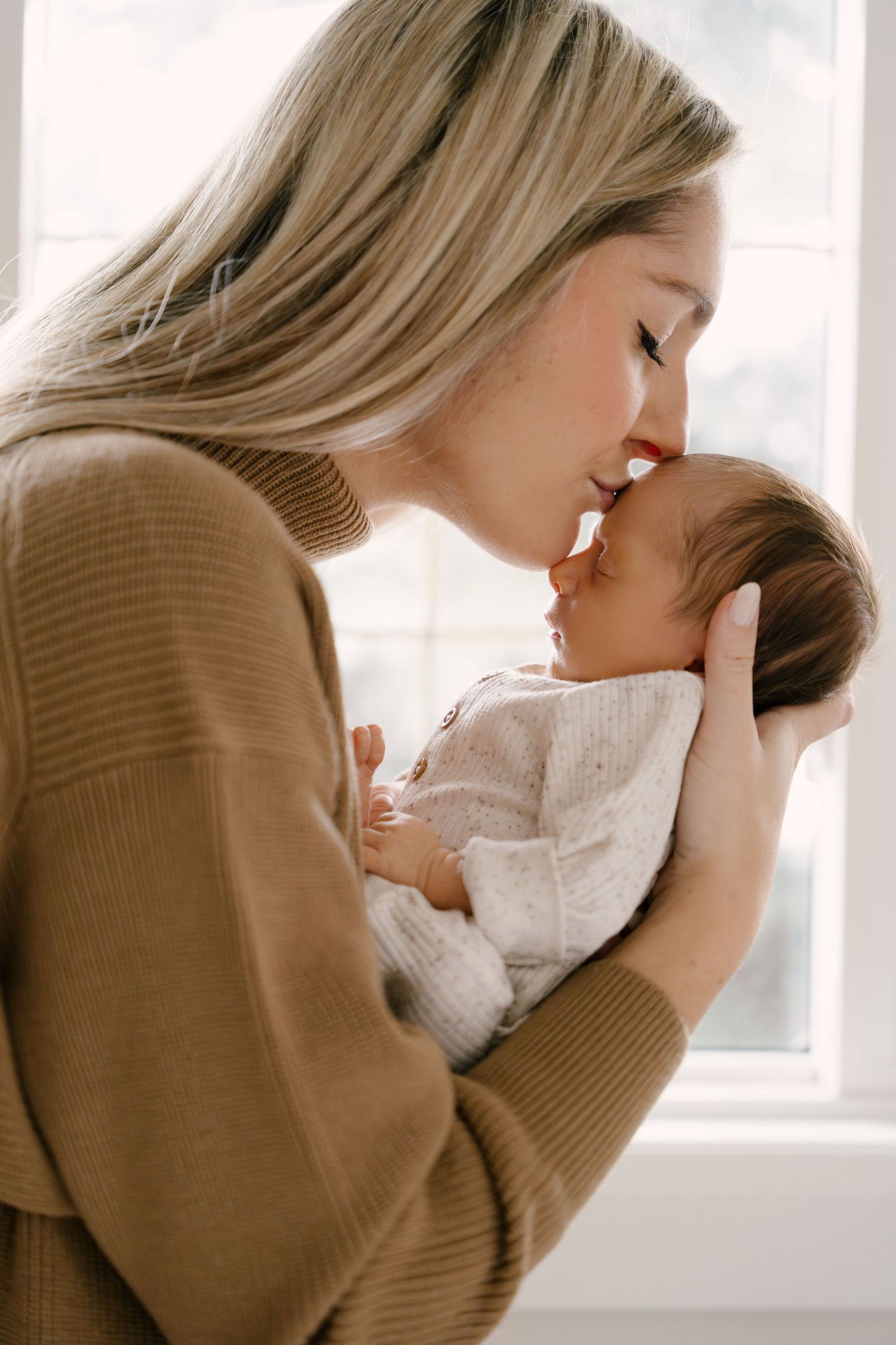 Tender moment of blonde mother touching noses with her newborn baby in white knit outfit.
