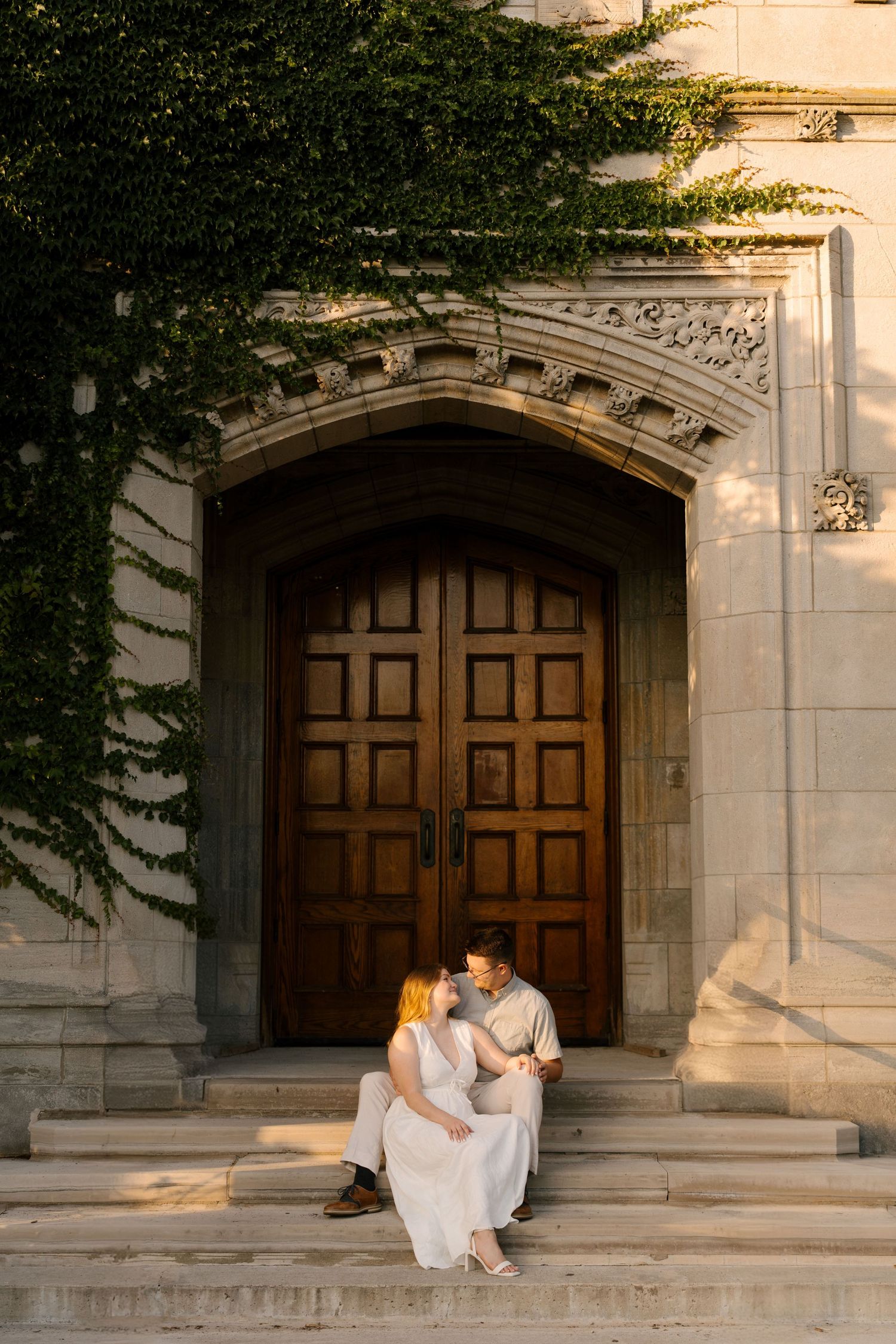 Couple sitting closely on steps of a stone building with ivy and large wooden doors at sunset.