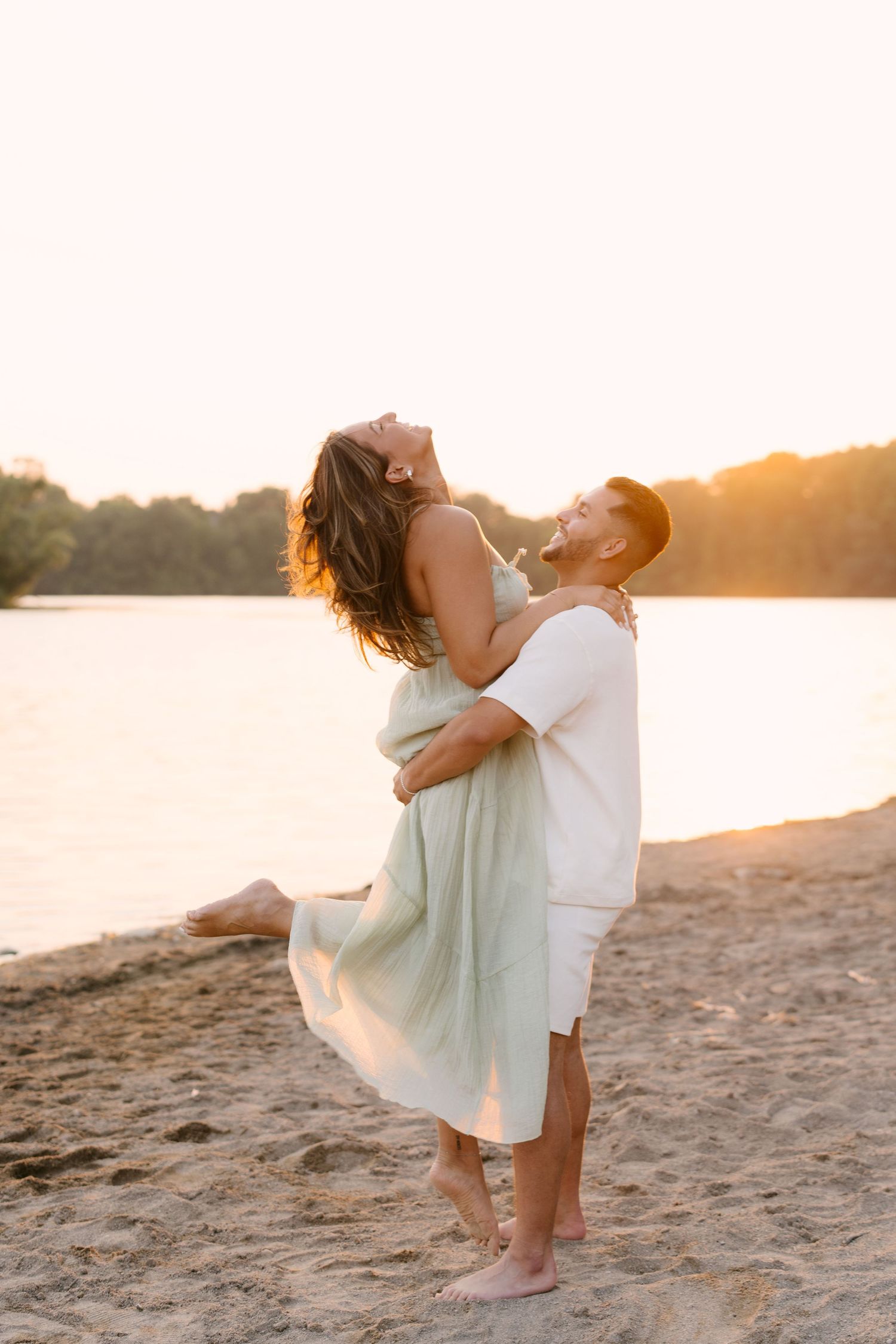 Couple embracing on sandy beach at golden sunset, man lifting woman in mint dress as she laughs joyfully.