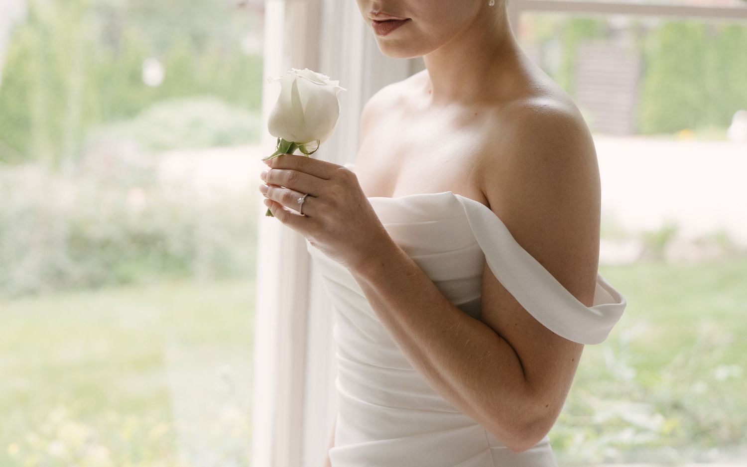 Bride in strapless white wedding gown holding a white rose near a sunlit window.