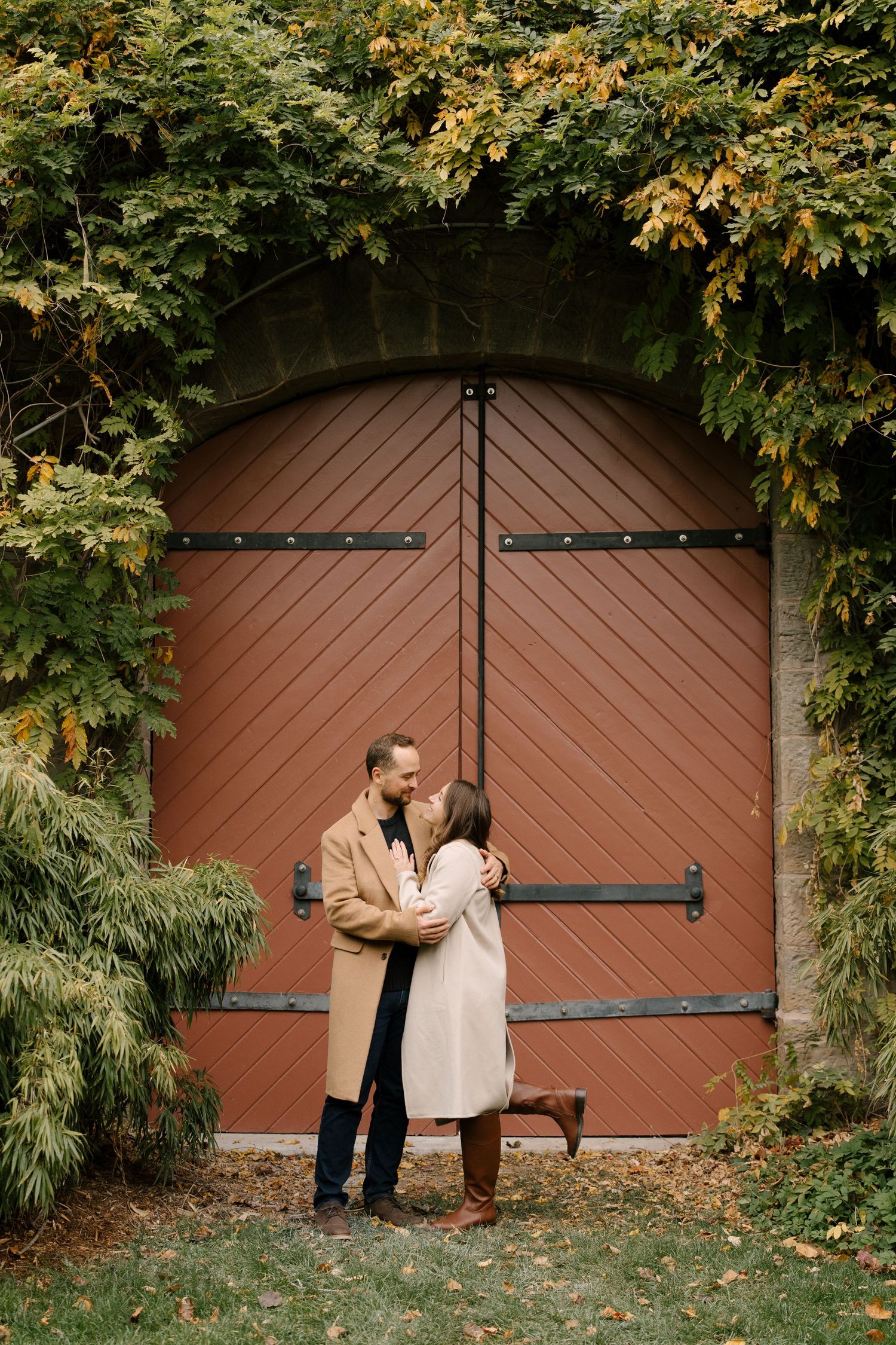 Couple kissing in front of a rustic red wooden door framed by lush autumn ivy and greenery in a garden.