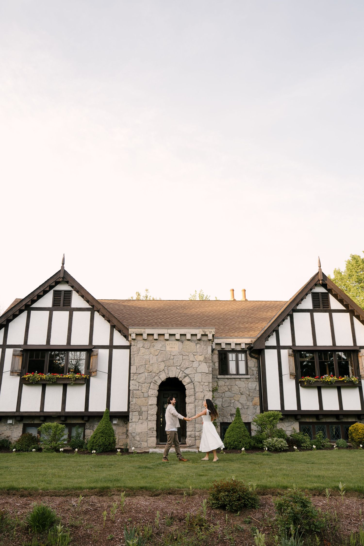 Couple holding hands while walking toward a charming Tudor-style manor house at dusk.