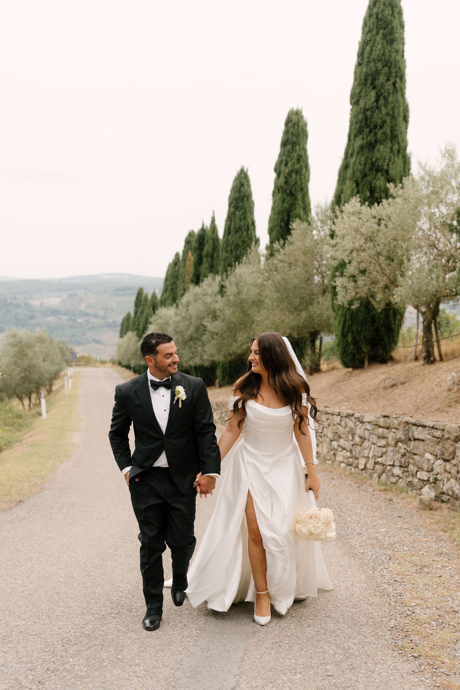 Bride and groom walk hand in hand along cypress-lined Tuscan road, she in slit gown with bouquet.