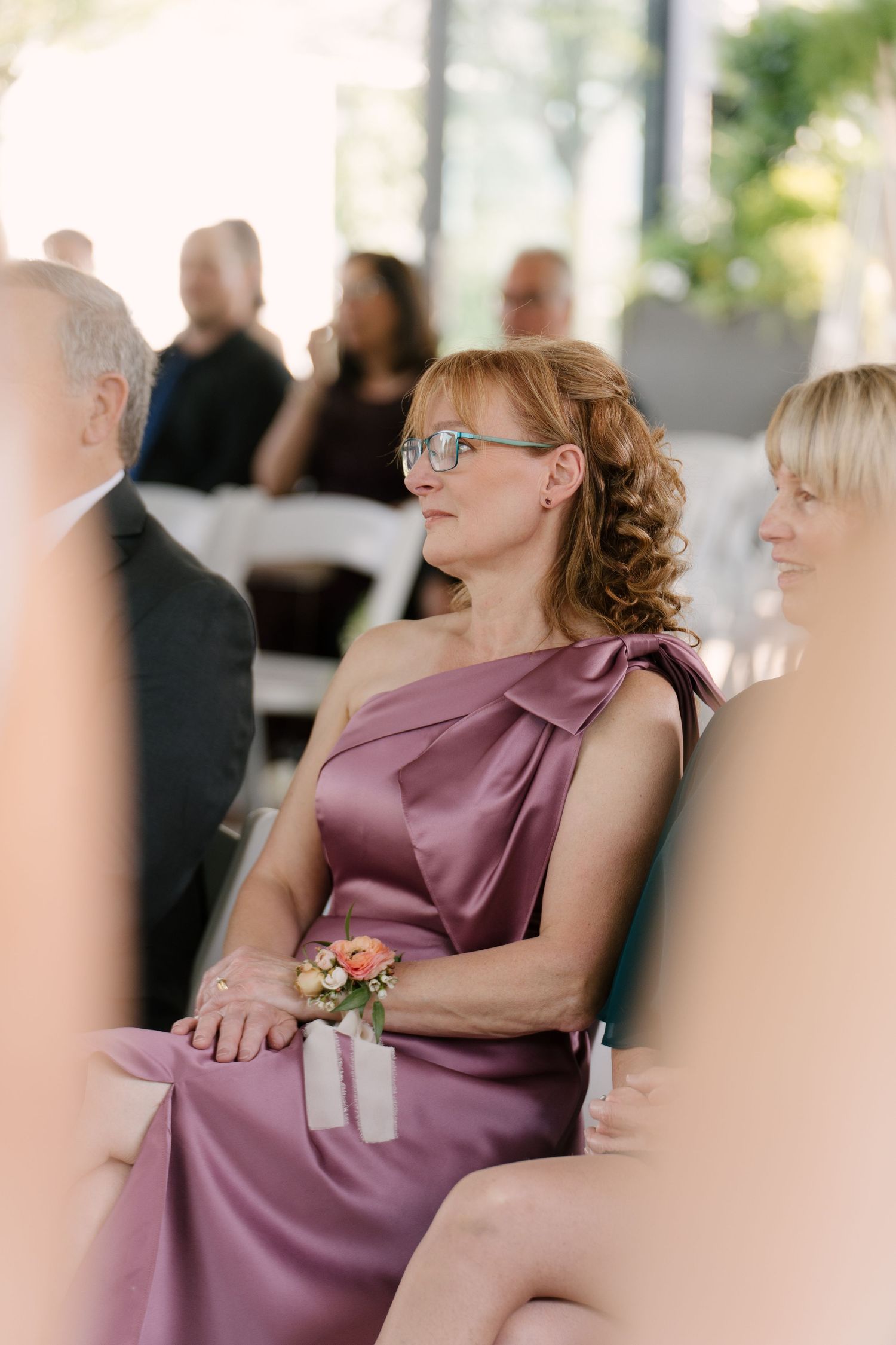 Wedding guest in one-shoulder dusty rose satin dress with wrist corsage seated during outdoor wedding ceremony.
