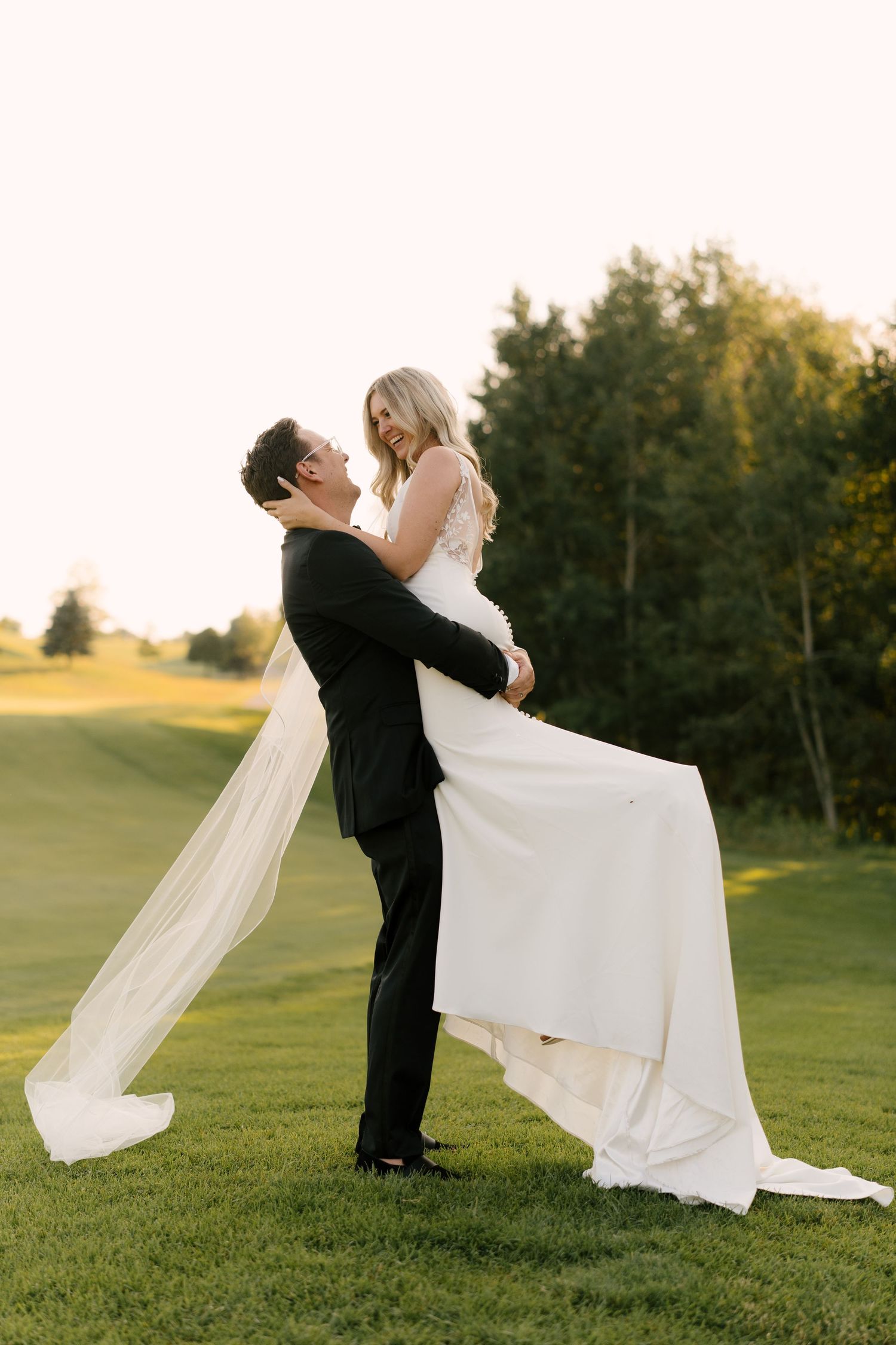 Groom lifting bride in white wedding gown on a lush green Whistle Bear golf course at sunset.