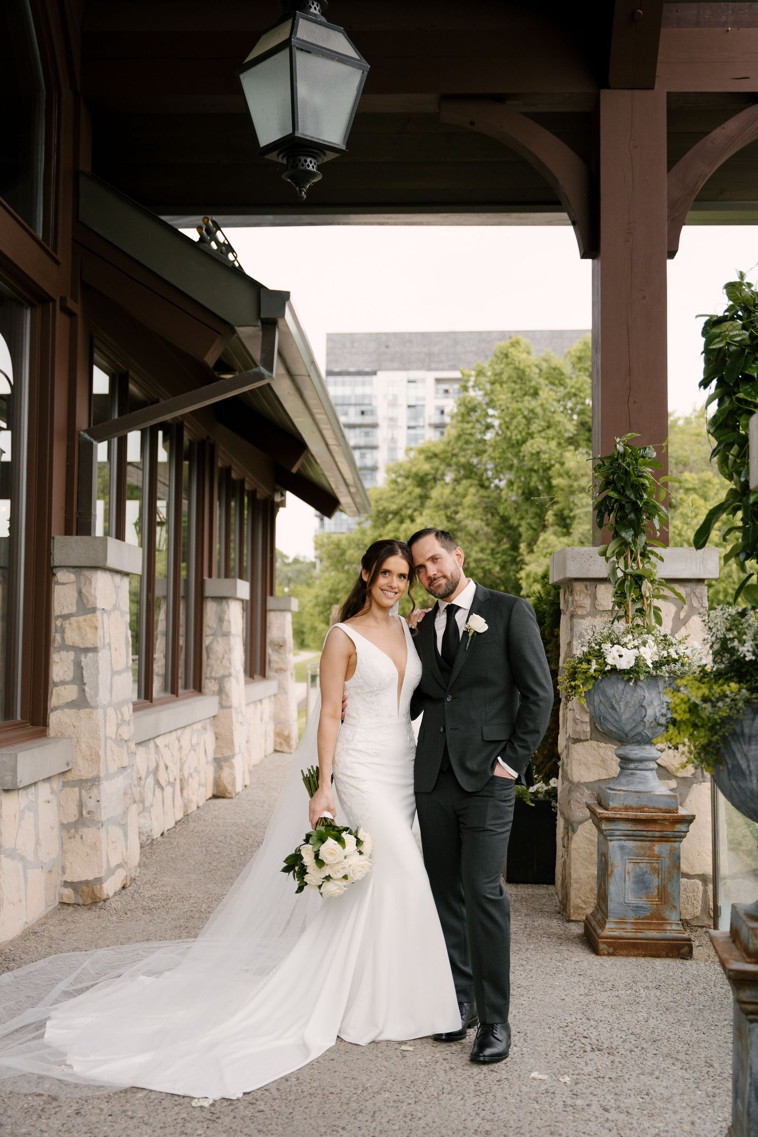 Bride and groom posing elegantly under a covered stone venue walkway with white floral bouquet.