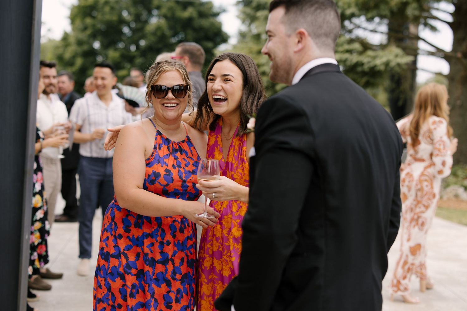 Wedding guests laughing and socializing outdoors, two women in colourful floral dresses holding wine.