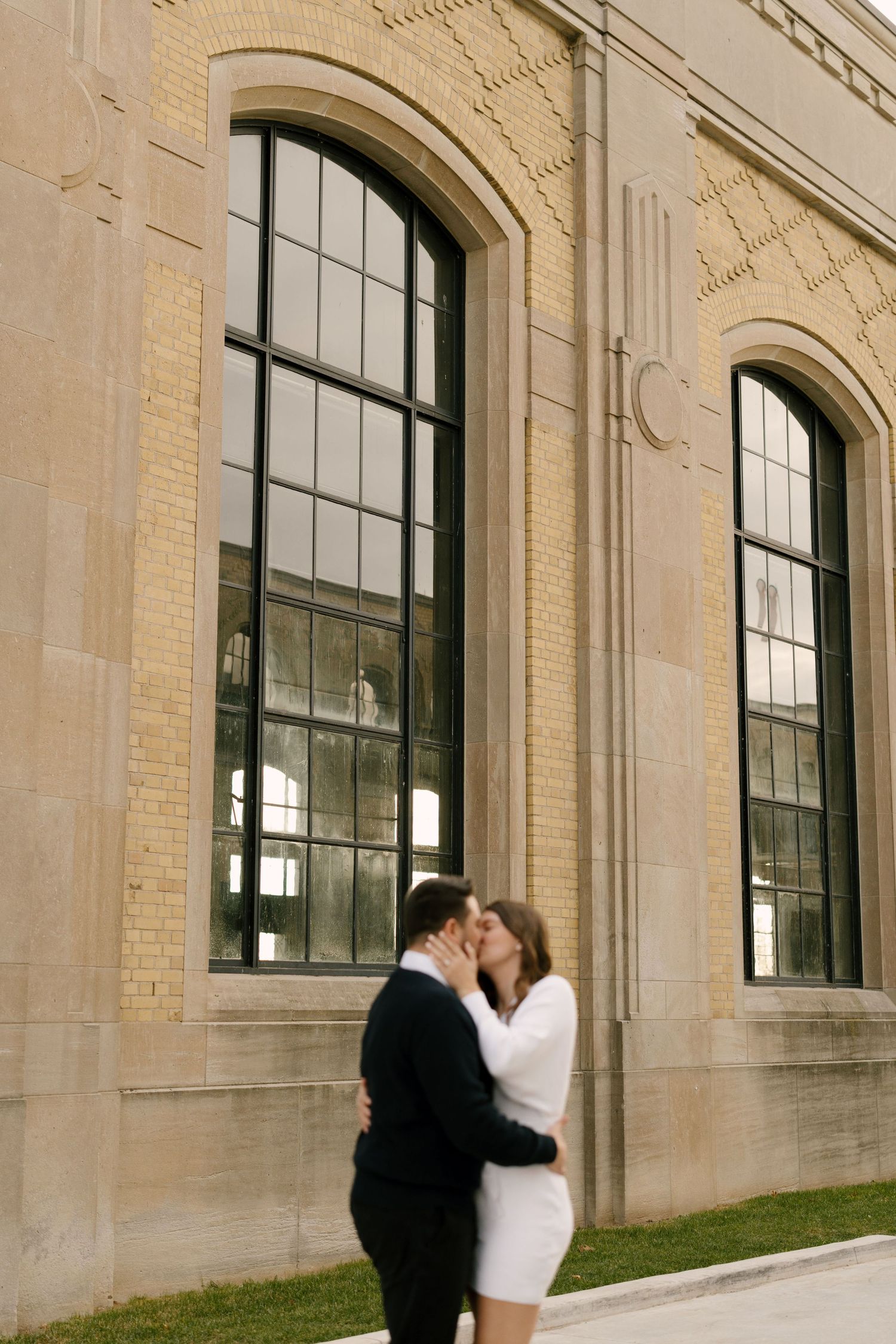 Couple sharing a kiss outside a grand historic brick building with tall arched windows.