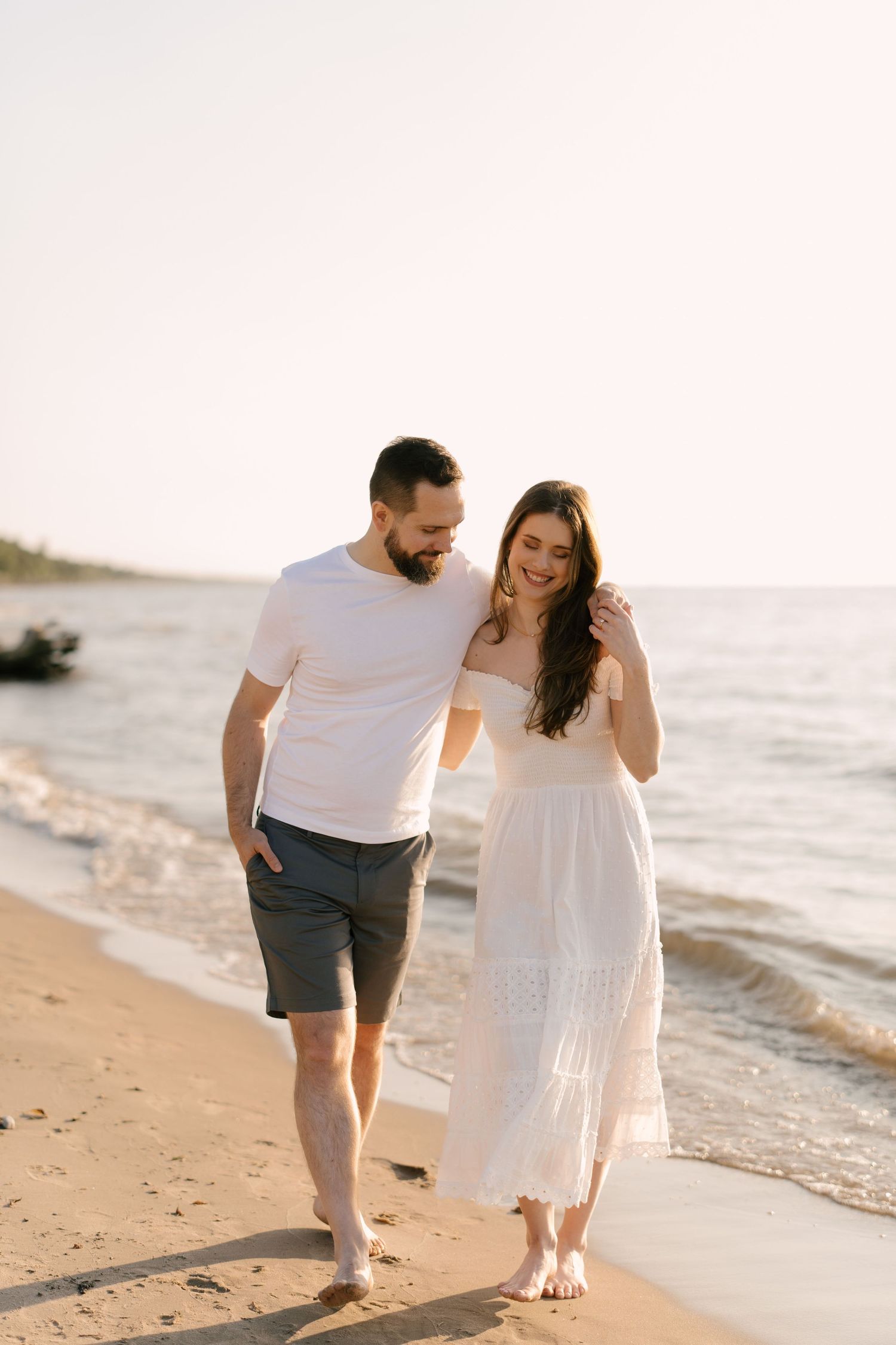 Romantic couple walking barefoot along a sandy beach at sunset, woman in flowing white dress.