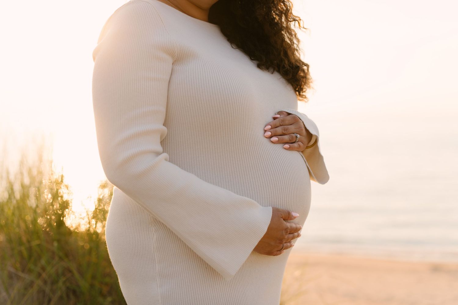 Close-up of pregnant woman in white dress cradling baby bump at golden hour beach.