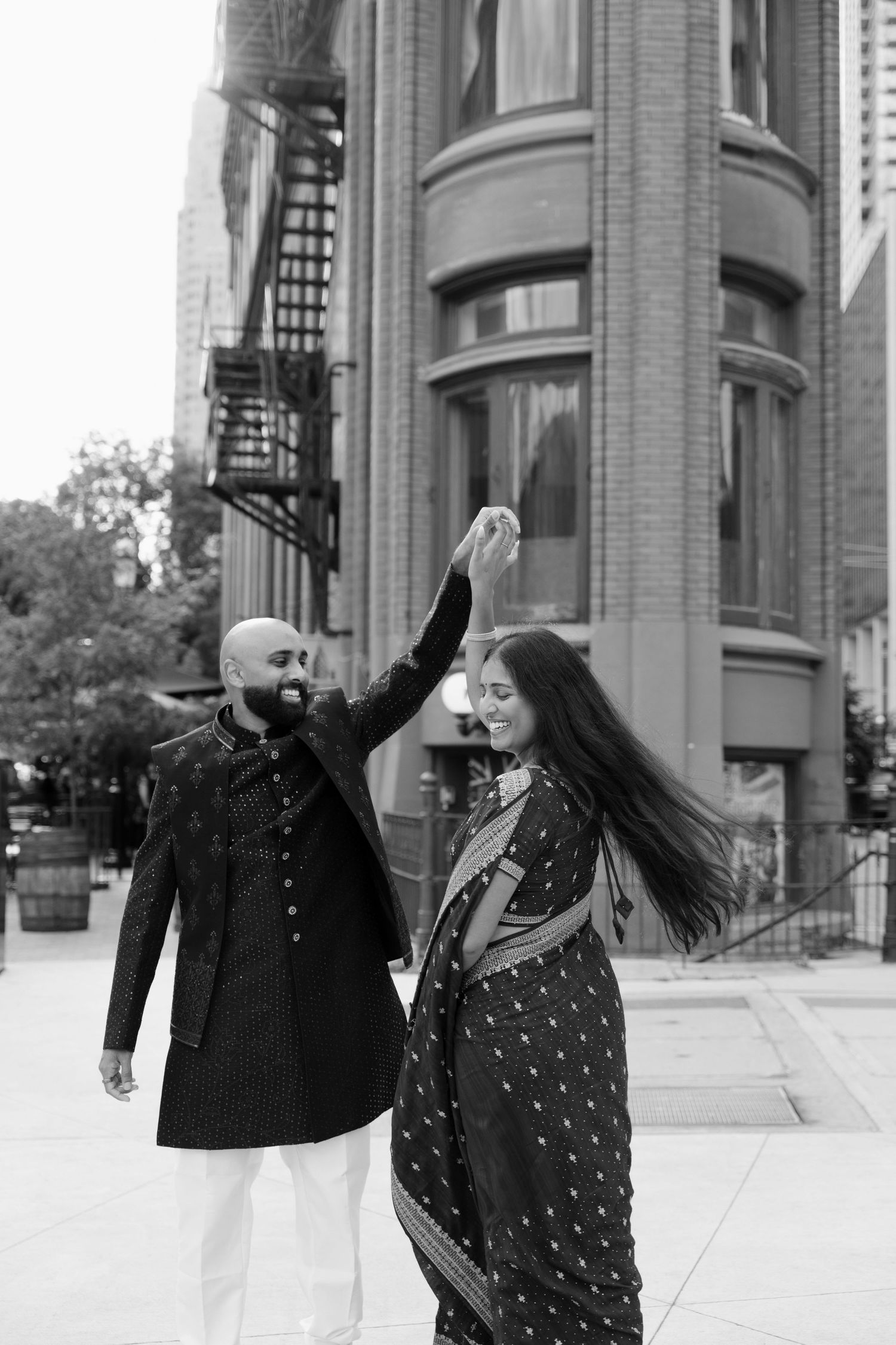 Black and white photo of Indian couple dancing in traditional attire near historic building.
