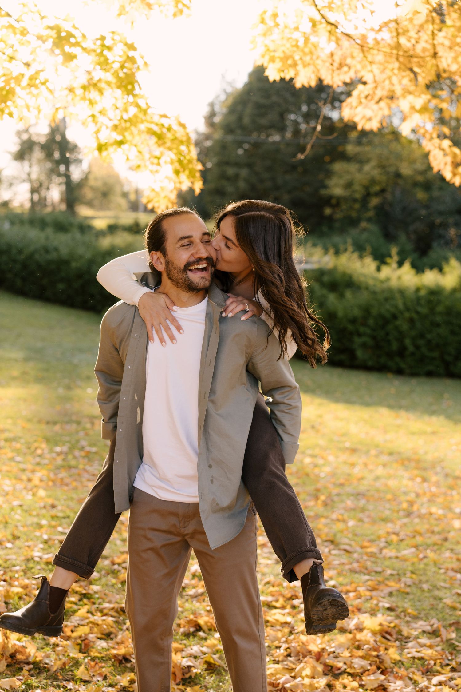 Woman kissing mans cheek as he laughs while giving her a piggyback in autumn park.