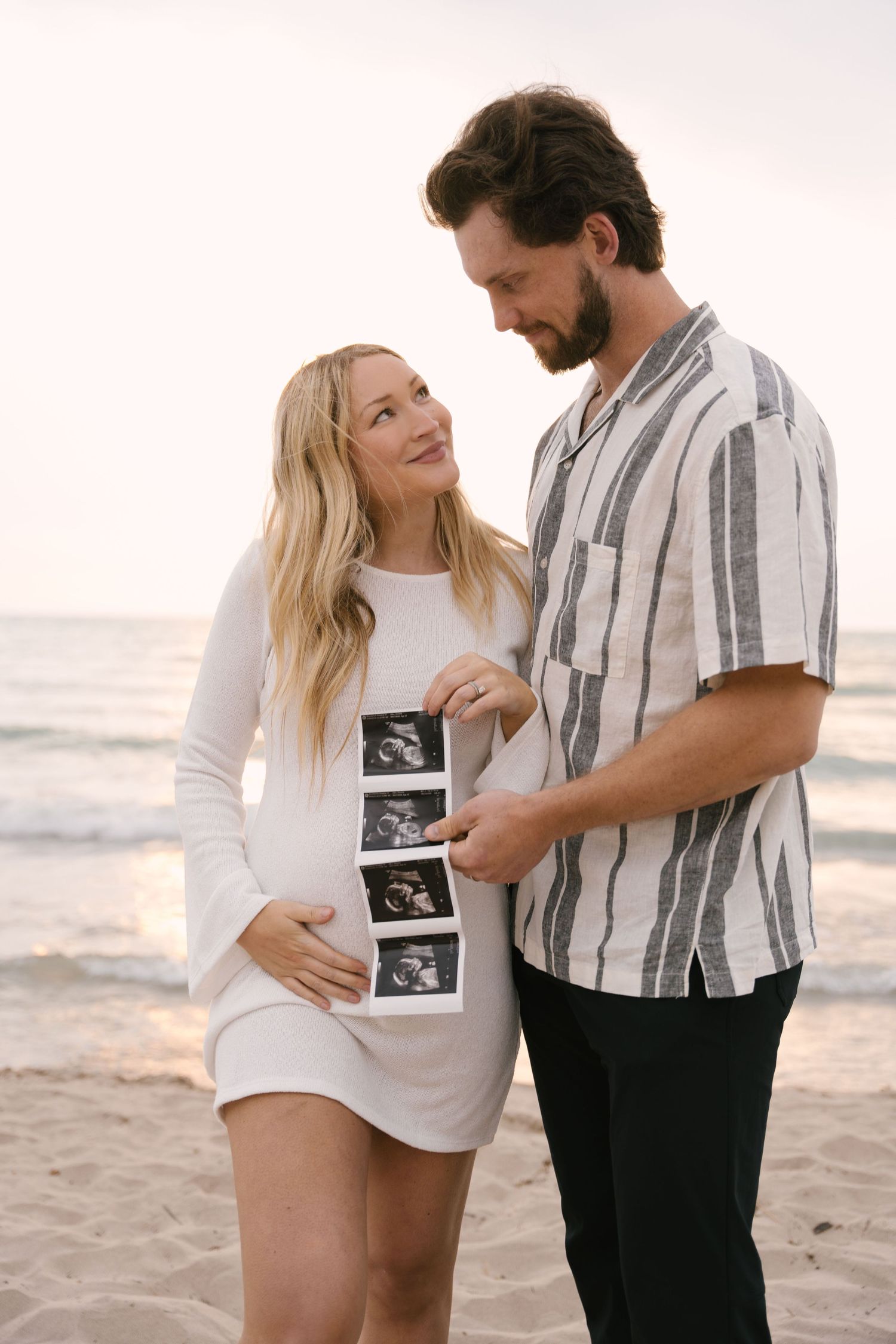 Expecting couple holding ultrasound photos together on sandy beach at dusk.