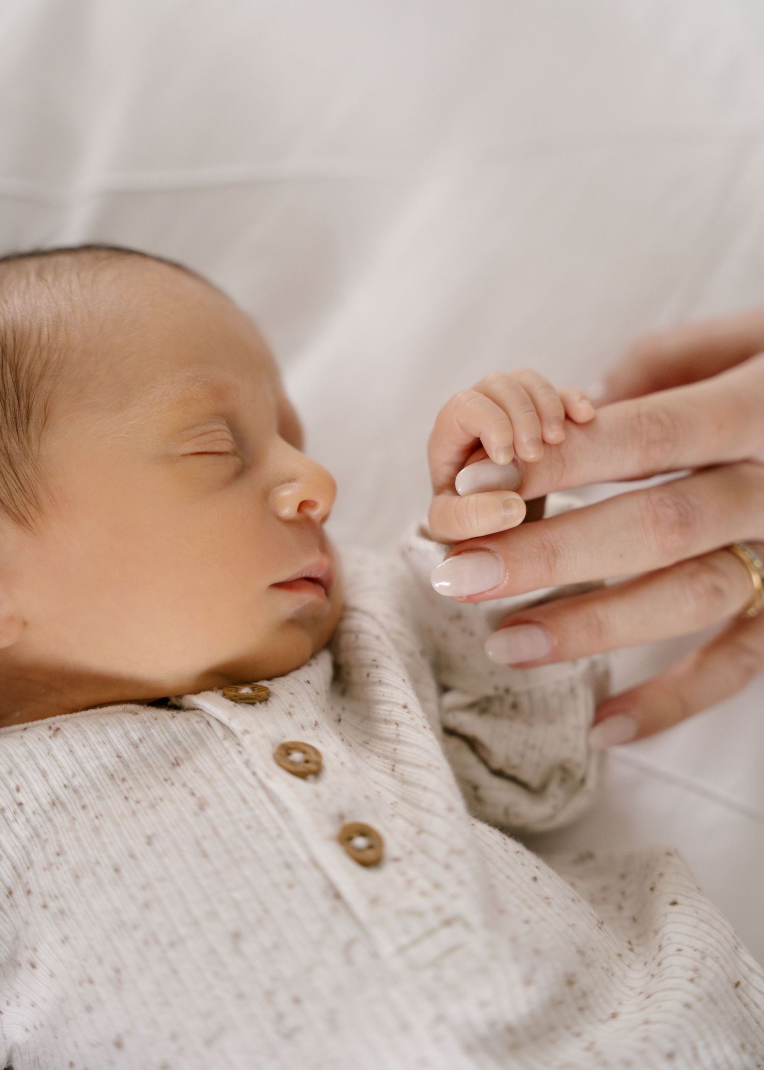 Sleeping newborn baby in cream knit outfit gently holding mothers finger with tiny hand.