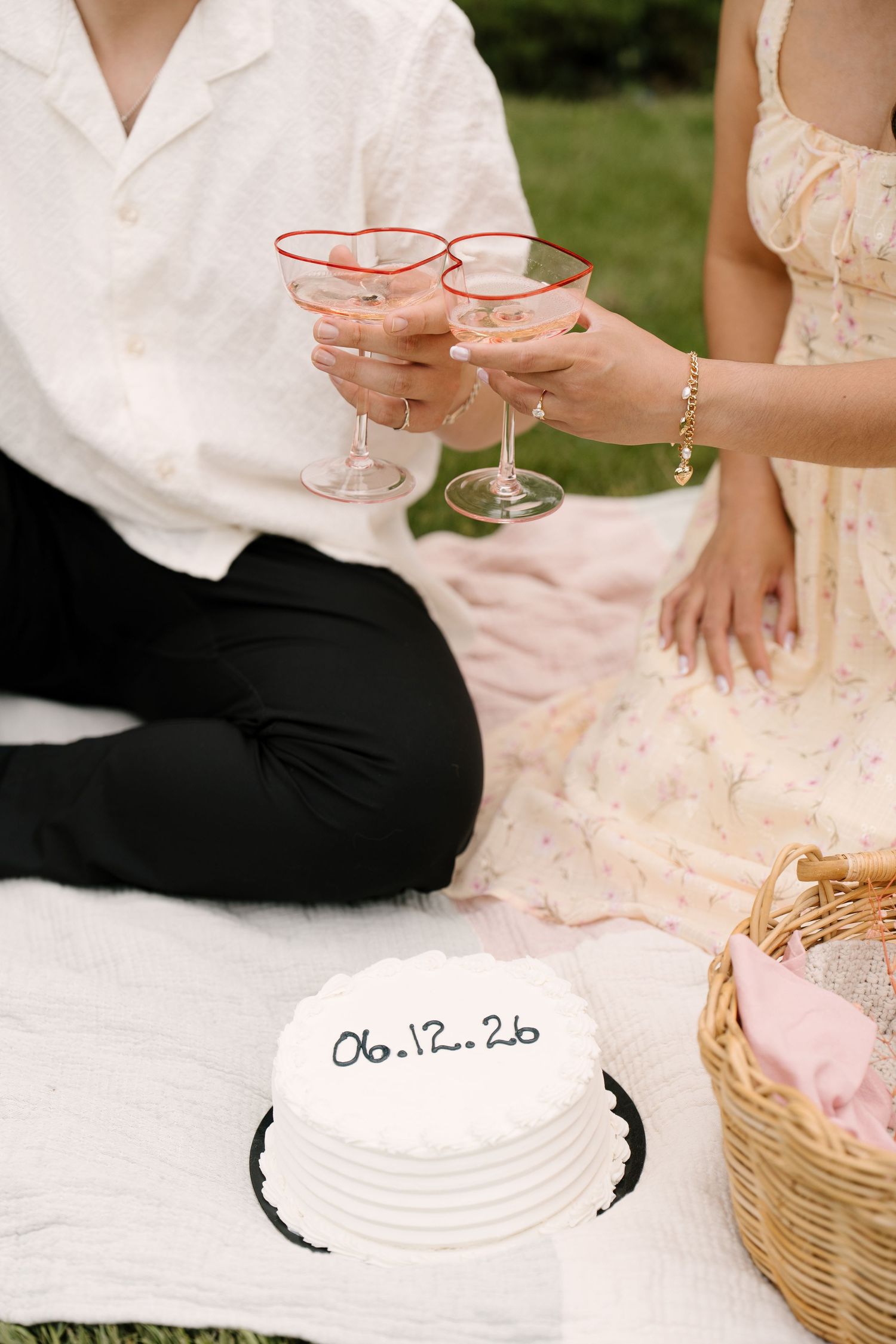 Couple clinking heart-rimmed champagne glasses beside a white cake reading 06.12.26 outdoors.