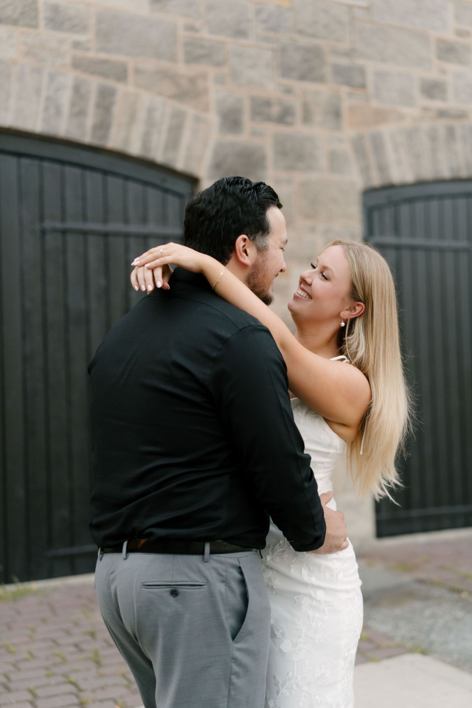Elegant couple embracing near stone building, woman in white lace dress and man in black shirt.