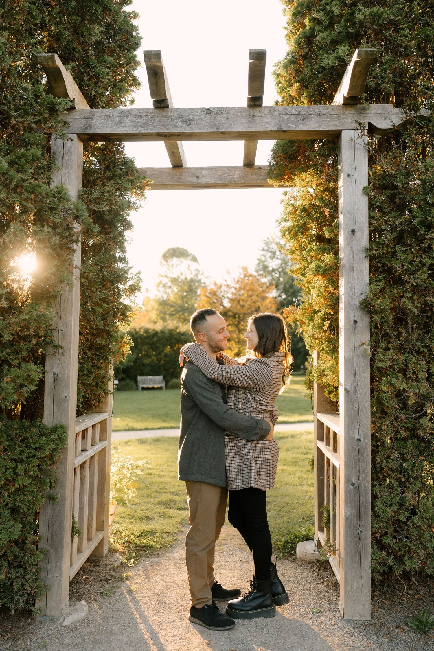 Couple embracing under wooden garden arbor surrounded by lush greenery in golden sunset light.